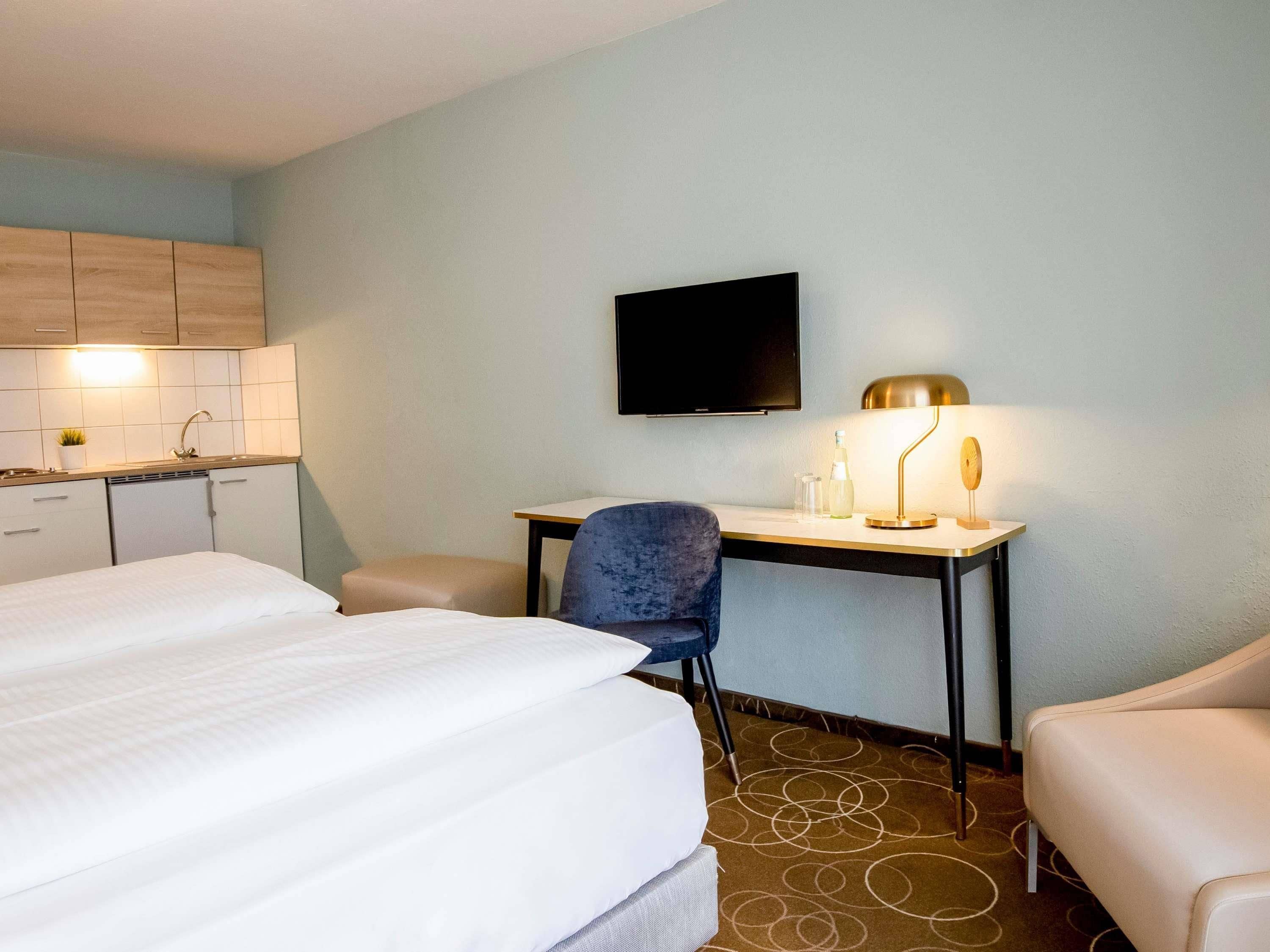 Ein Hotelzimmer mit Bett, Schreibtisch, Stuhl, Fernseher und einer kleinen Küchenzeile.