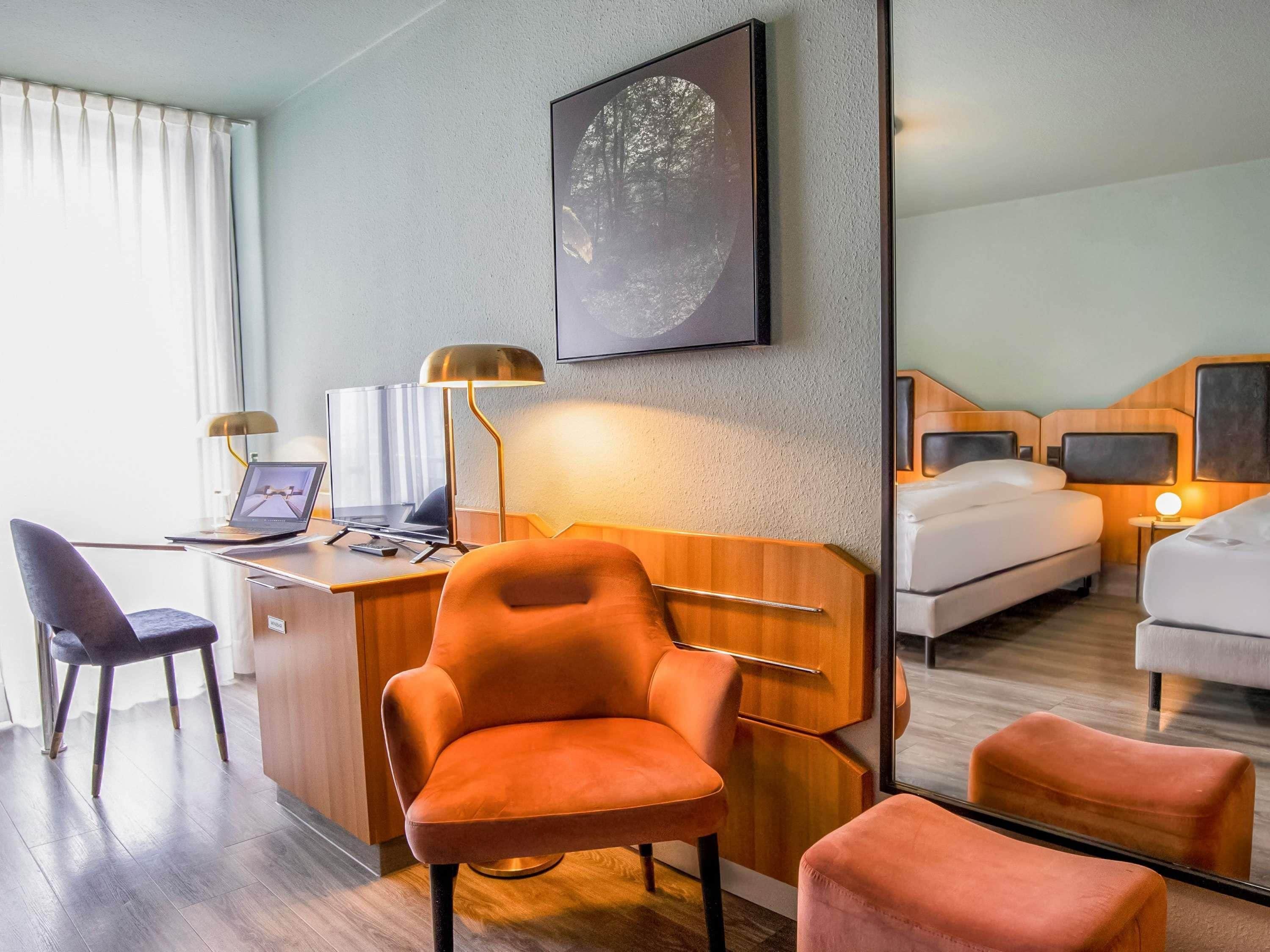 A hotel room with a desk, laptop, orange chair, bed, bedside table, lamp, and a mirror.