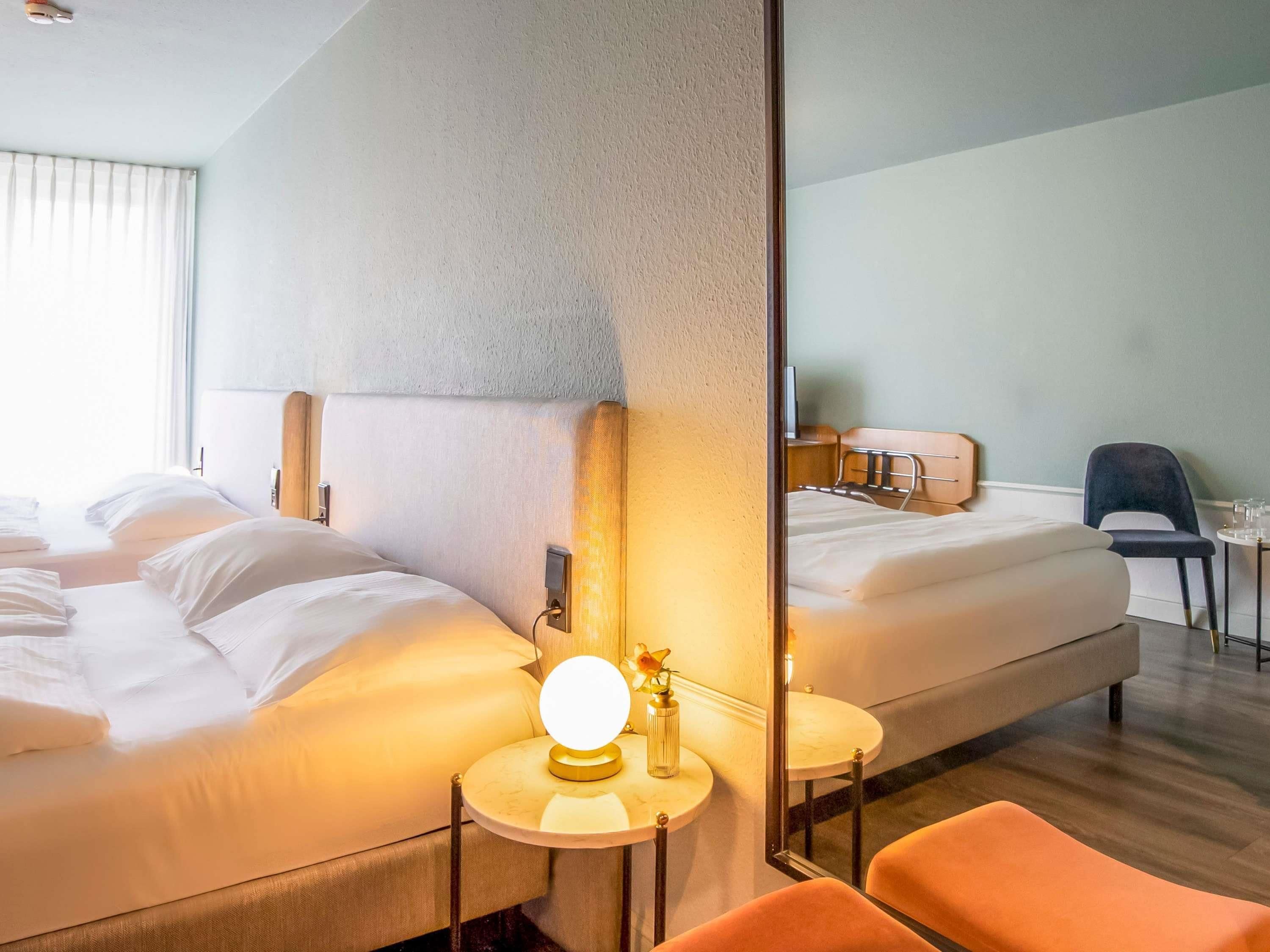 A hotel room with two beds, a round table with a lamp, a chair, and a large mirror.