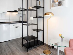 A modern kitchen with white cabinets, a black metal shelving unit, a glass table with a gold lamp, and a pink chair. - Mercure Hotel Pforzheim (Pforzheim)