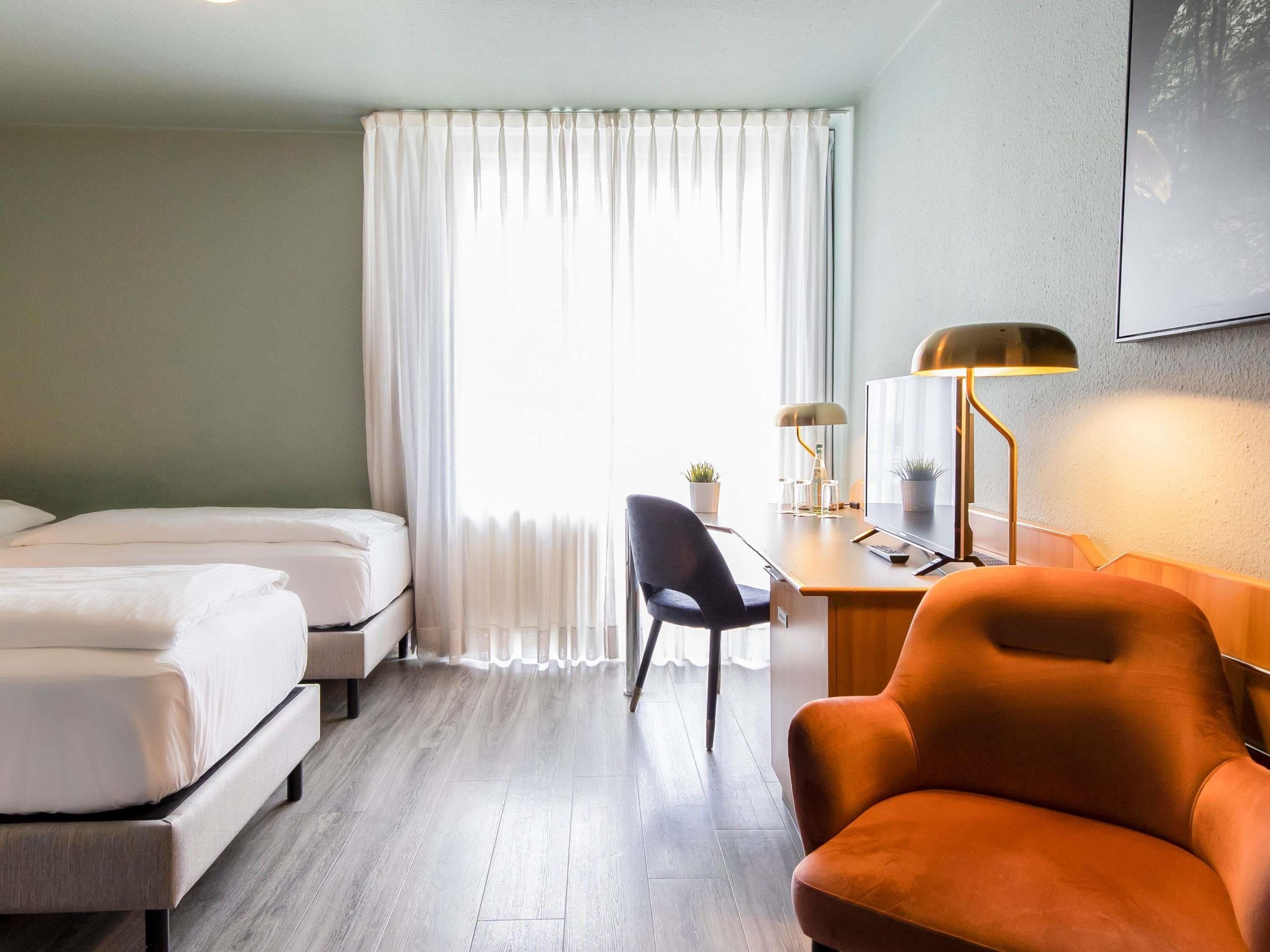 A hotel room with two beds, a desk with a lamp, a chair, and a large window with curtains.