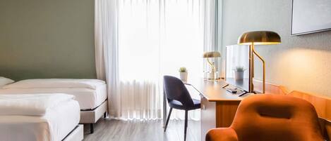 A hotel room with two beds, a desk with a lamp, a chair, and a large window with curtains.