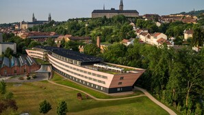 Un hôtel moderne doté d’un toit incurvé original, entouré d’une végétation luxuriante et bordé de bâtiments historiques en arrière-plan.
