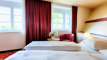 Ein Hotelzimmer mit zwei Betten, einem roten Sessel, einem kleinen Tisch mit Blumenvase und einem Fenster mit roten Vorhängen.