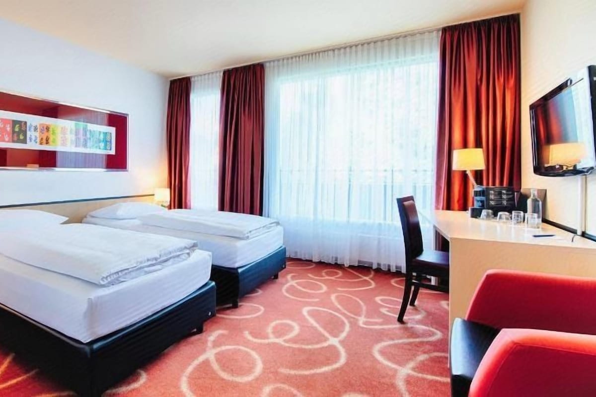 Una habitación de hotel con dos camas, un escritorio, una silla, un televisor y una ventana con cortinas.