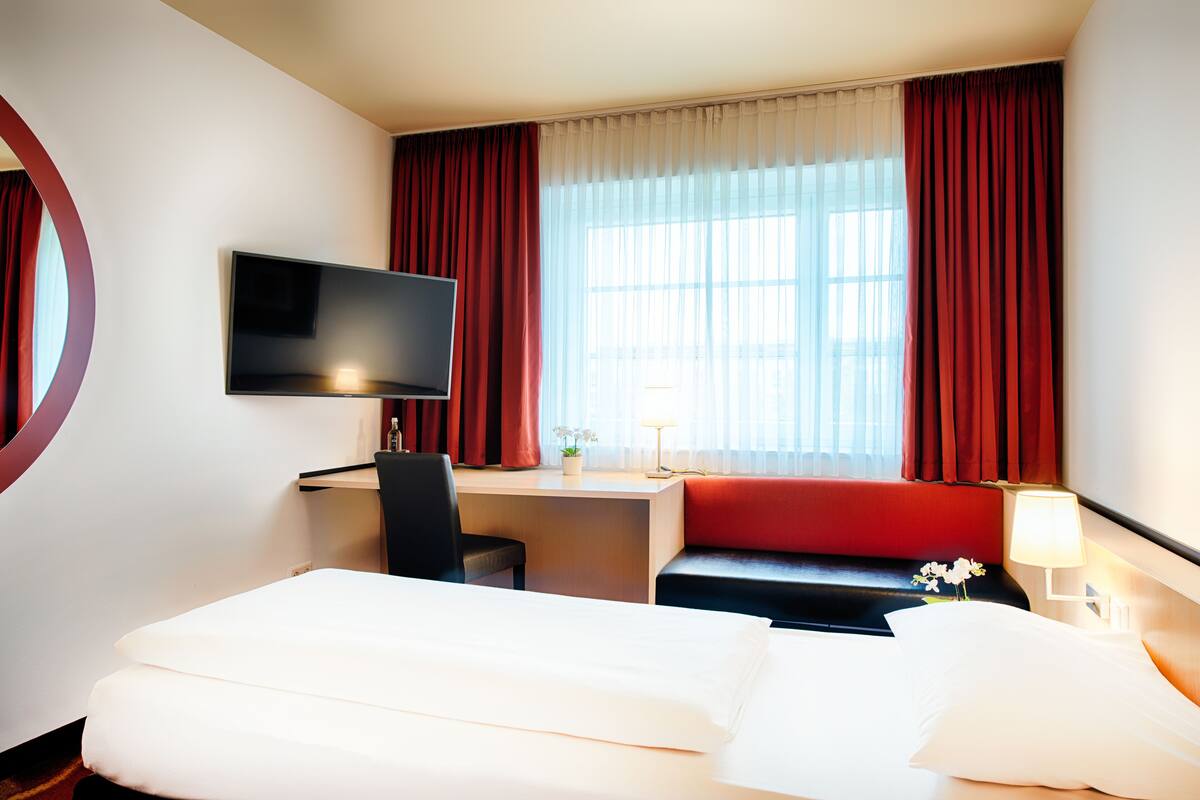 Habitación de hotel con cama, escritorio, silla, televisor, lámpara y ventana con cortinas.