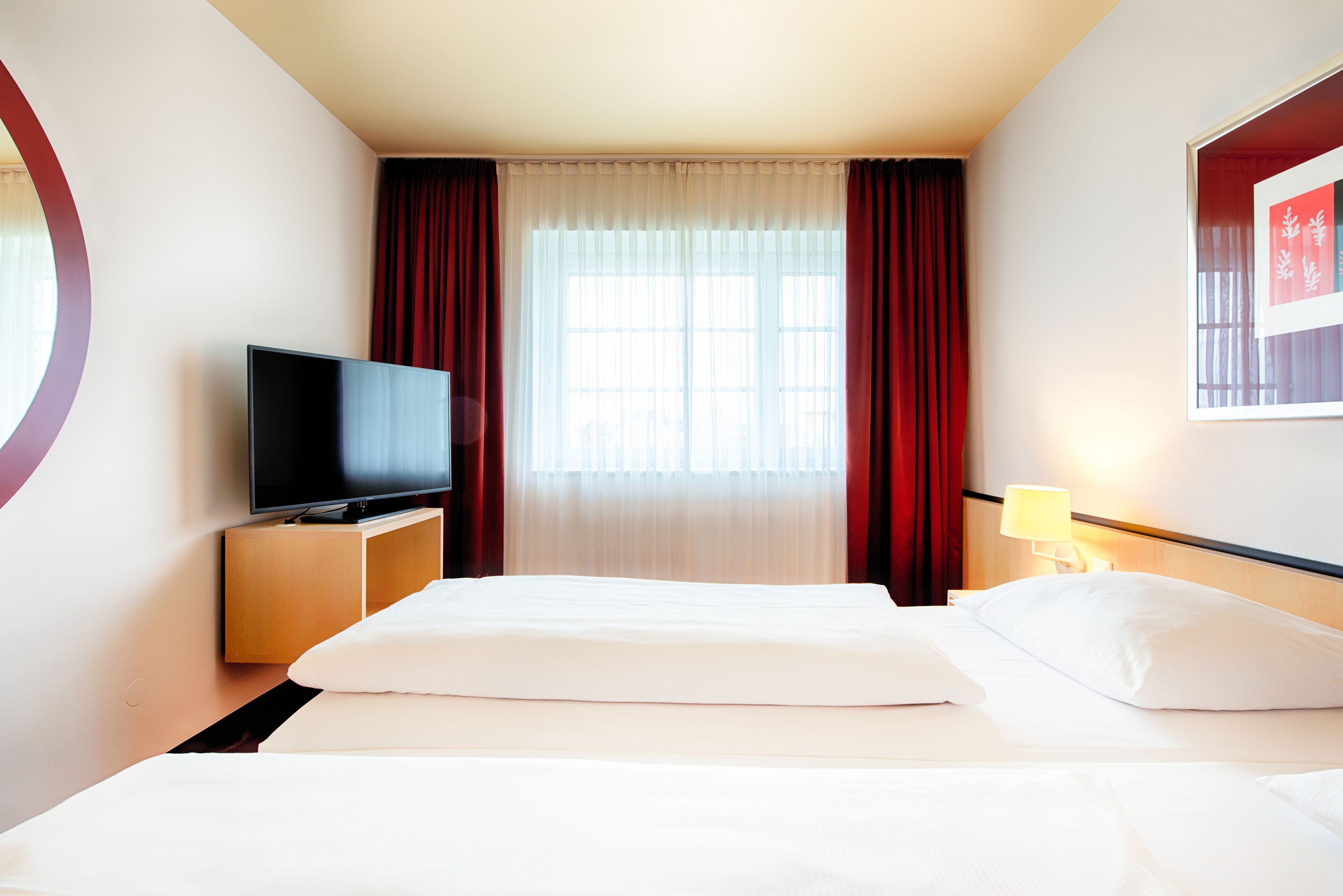 Une chambre d’hôtel avec un lit, un téléviseur posé sur un meuble en bois, une fenêtre avec des rideaux et une lampe fixée au mur.