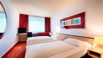 A hotel room with two beds, a desk, a TV, and a window with red curtains.