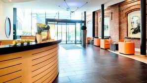 Un espace d’accueil moderne avec un comptoir en bois aux lignes courbes, un coin salon avec des fauteuils orange et un mur en briques orné d’une fresque.