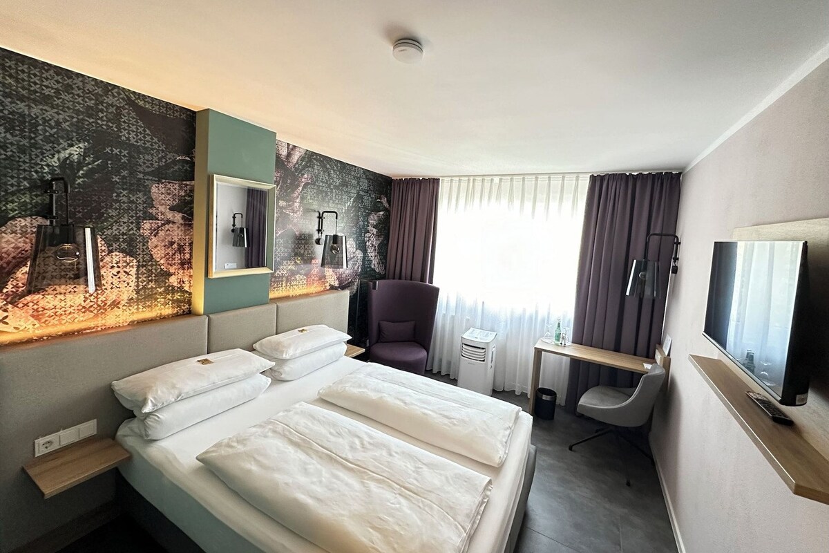 Habitación de hotel con cama, escritorio, silla, televisor y ventana con cortinas.
