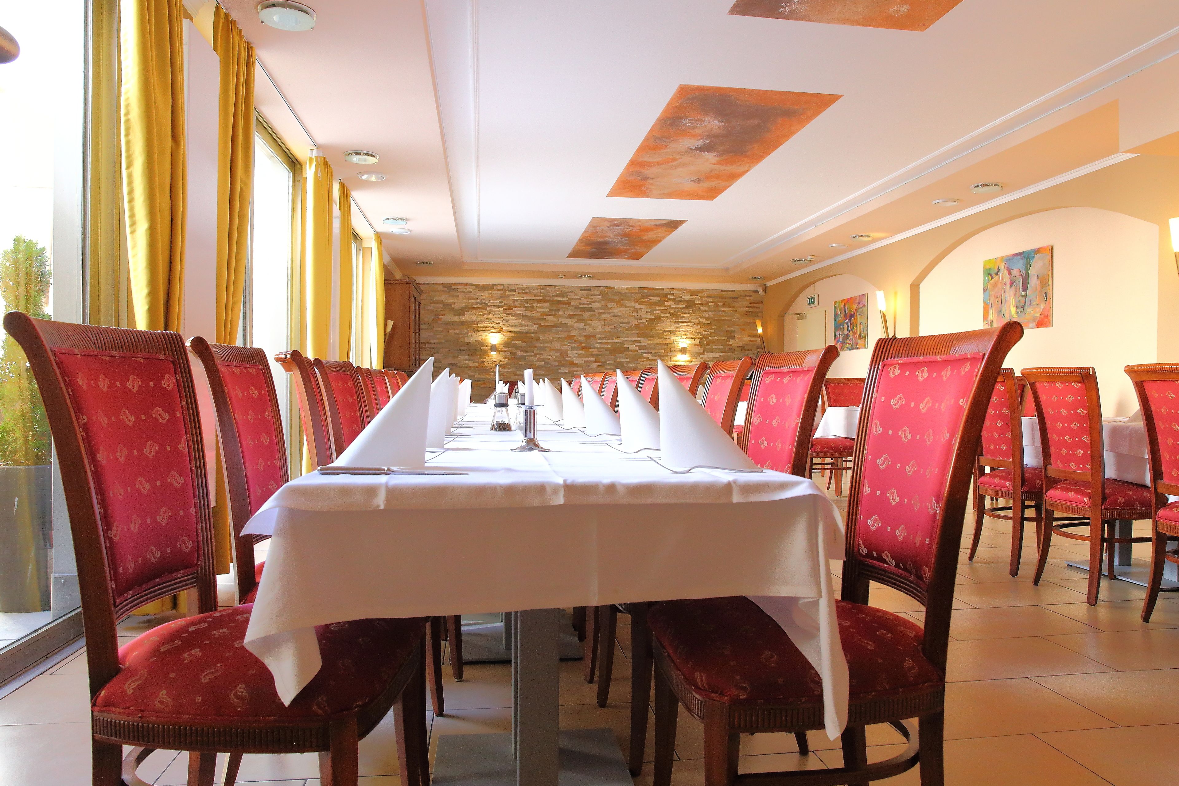 Une longue table à manger, avec des chaises à revêtement rouge, des nappes blanches et un mur en pierre en arrière-plan.