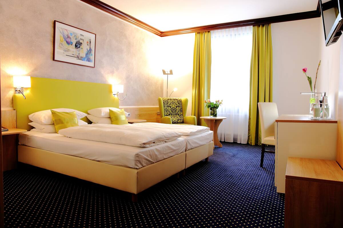 A hotel room with a bed, a desk, a chair, a small table, and a lamp.