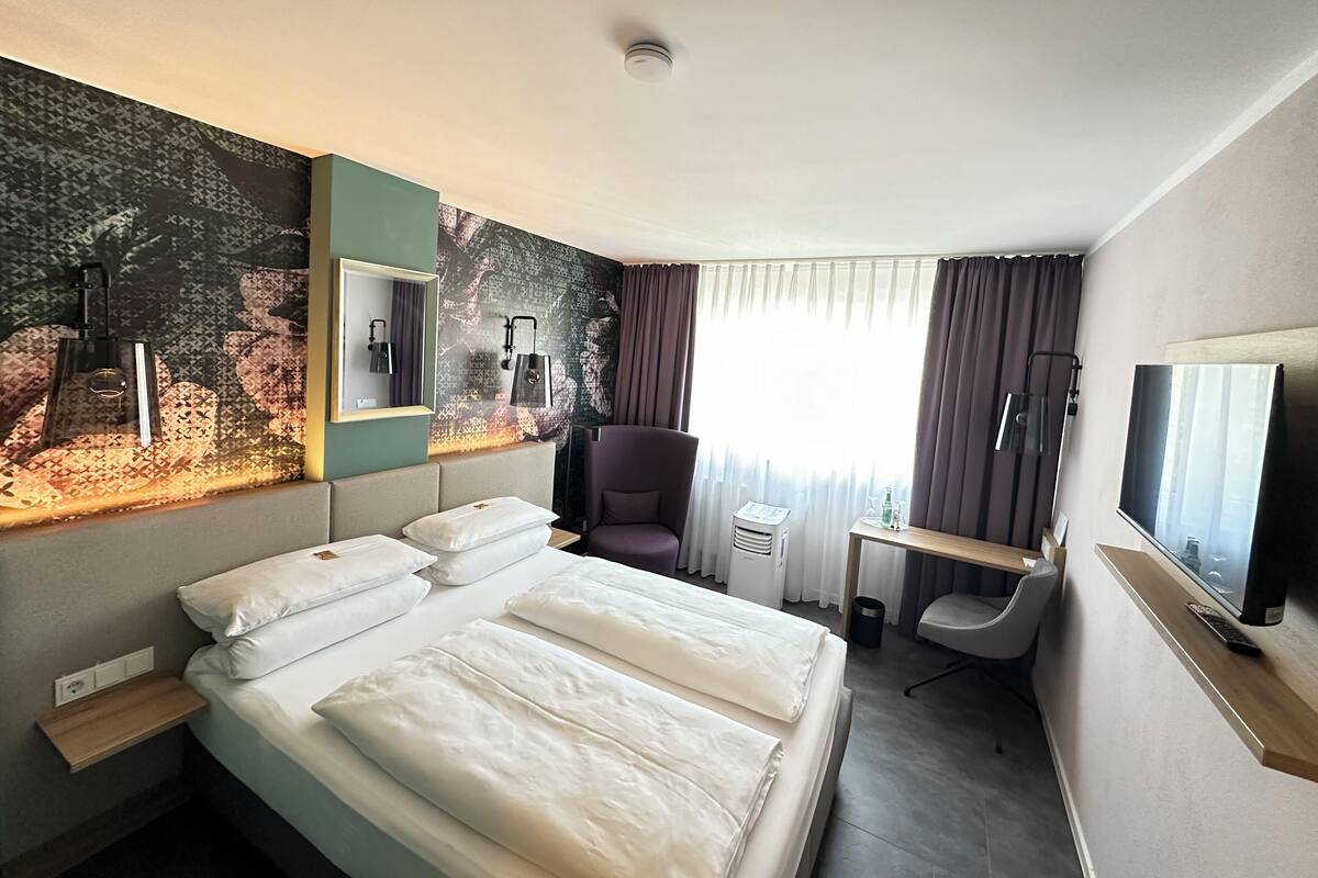 A hotel room with a bed, a desk, a chair, a TV, and a window with curtains.