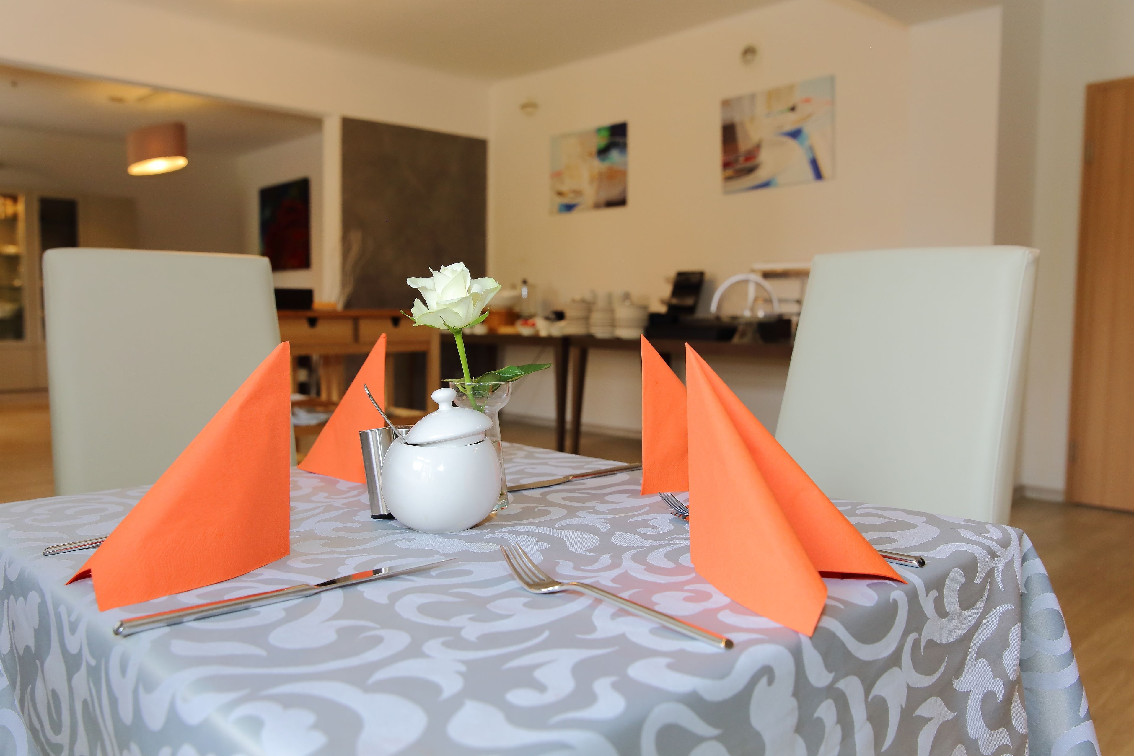 A dining table with a patterned tablecloth, orange napkins, a white vase with a single white flower, and silverware.