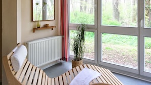 A room with a curved wooden bench, a mirror, a shelf with a vase, and large windows with a view of trees. - Hotel Haus Duden (Wesel)