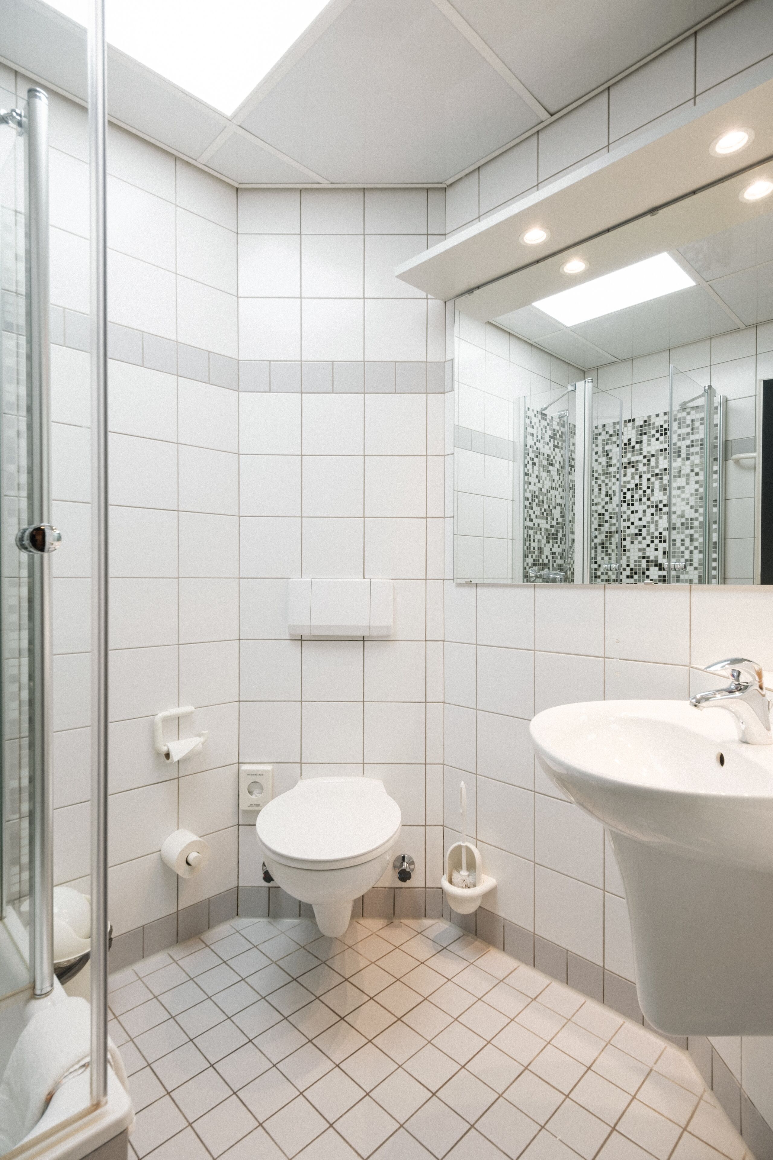 A modern bathroom with white tiles, a glass shower enclosure, a toilet, and a sink with a mirror above it.