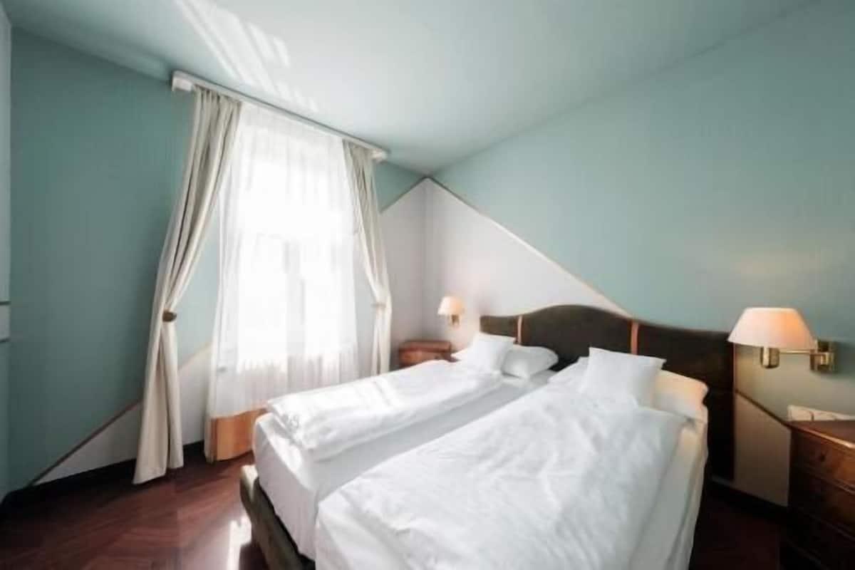 Ein Hotelzimmer mit zwei Betten, einem Fenster mit Vorhängen, einem Nachttisch mit Lampe und einer Wandlampe.