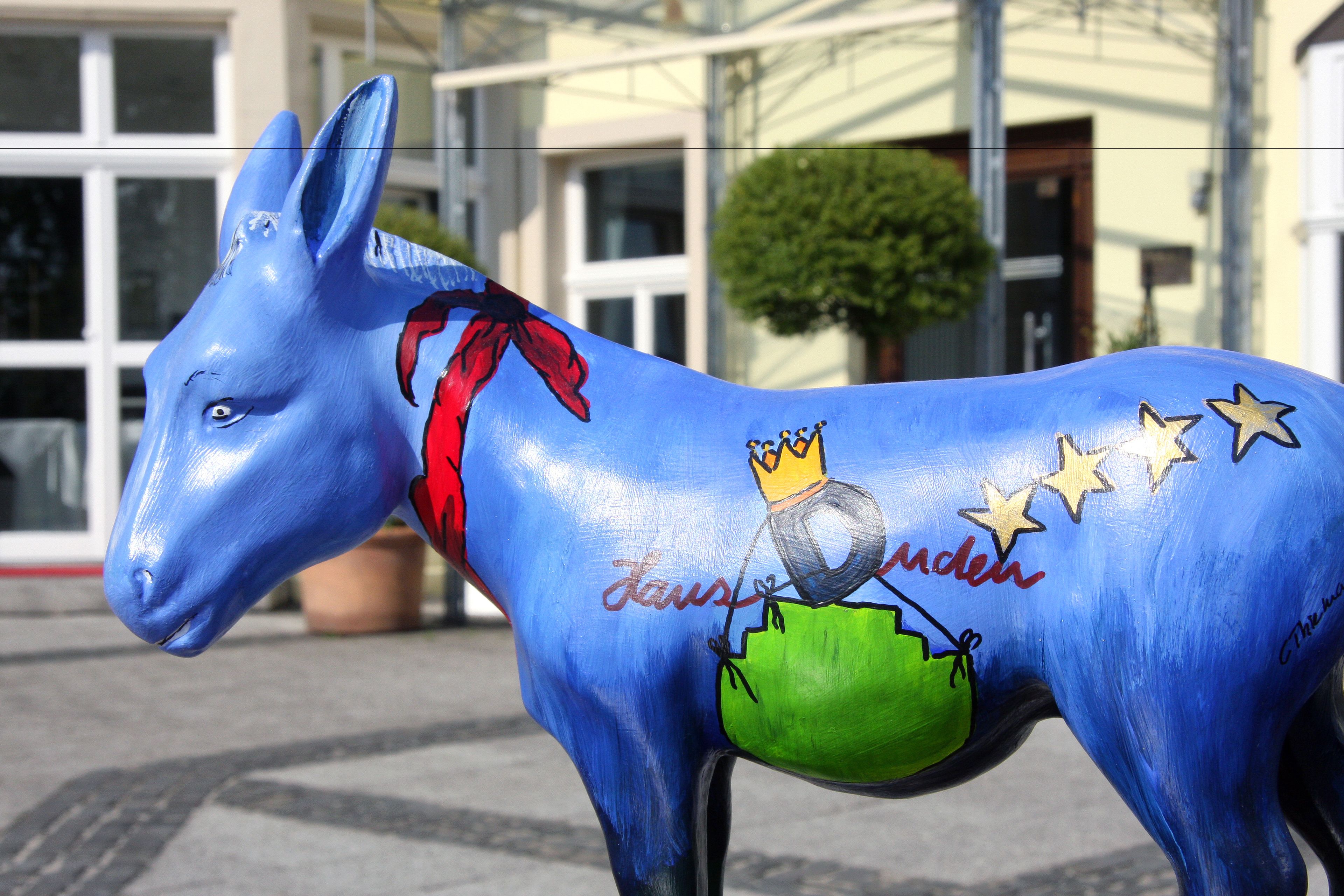 Una estatua de burro azul con una cinta roja y estrellas, ubicada frente a un edificio de grandes ventanales y una planta en maceta.