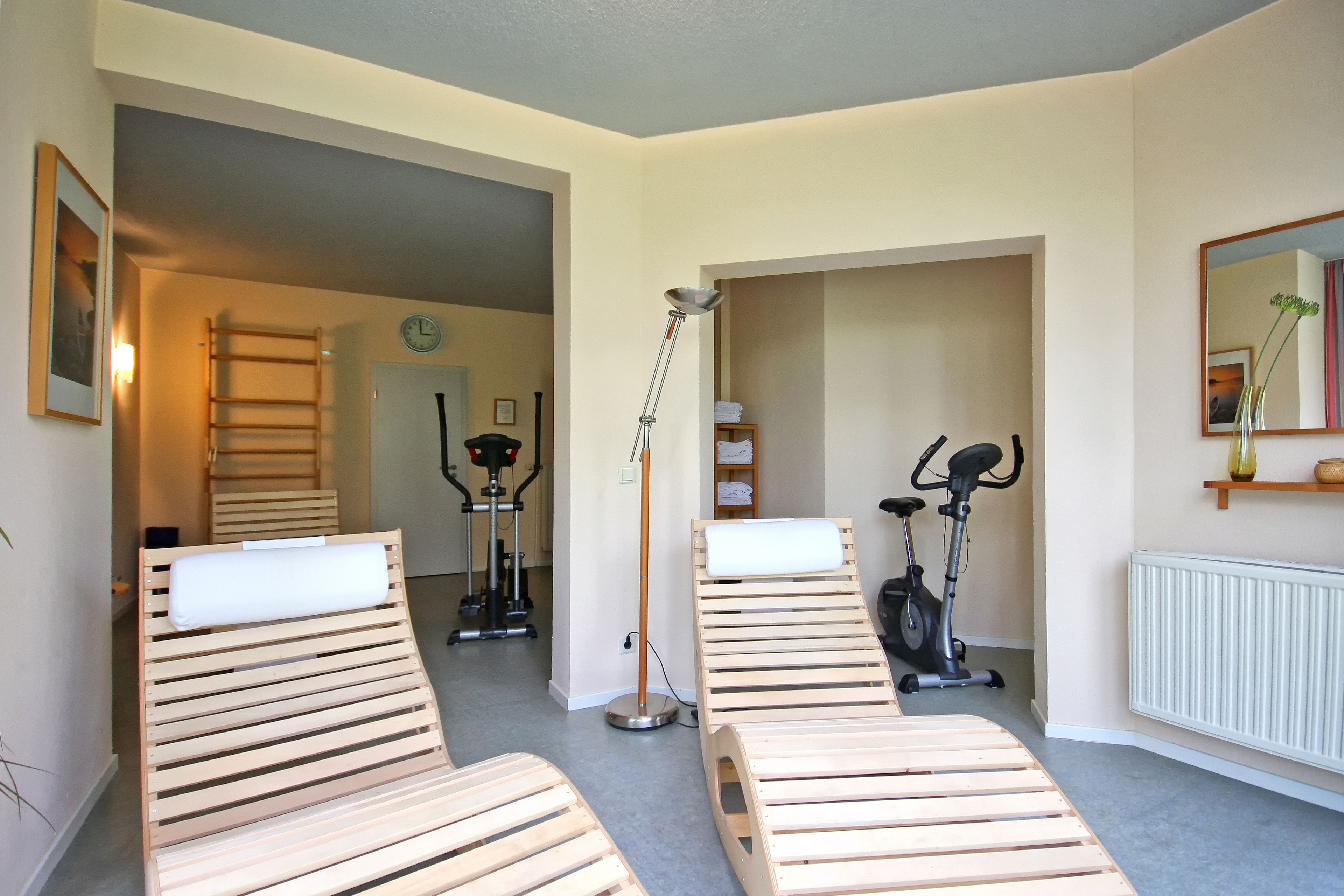 Una habitación con dos sillones de madera, una lámpara de pie, bicicletas estáticas, una escalera, un reloj y un radiador.