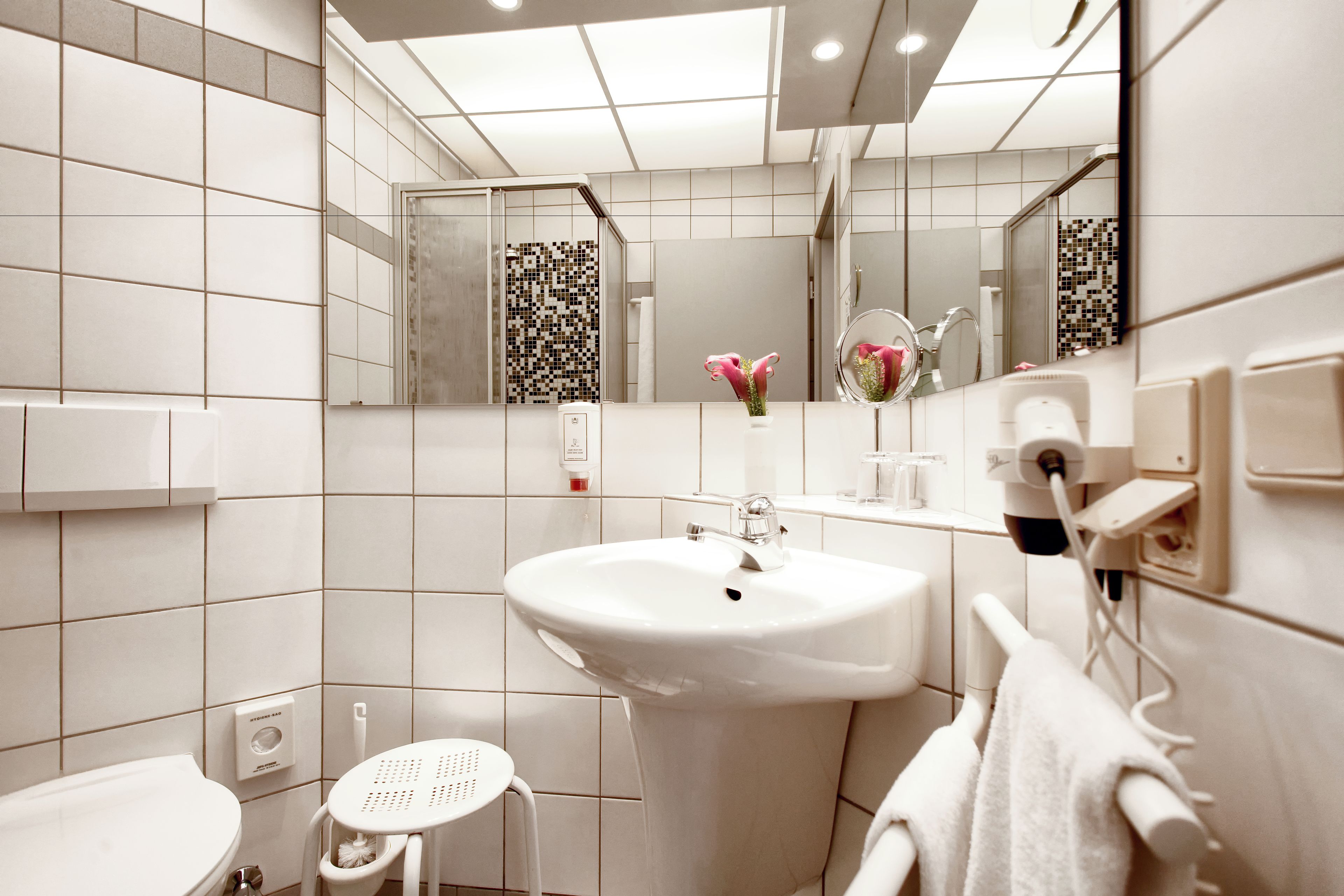 A modern bathroom with a white sink, toilet, and bidet. It includes a mirror, a towel rack, and a small stool.