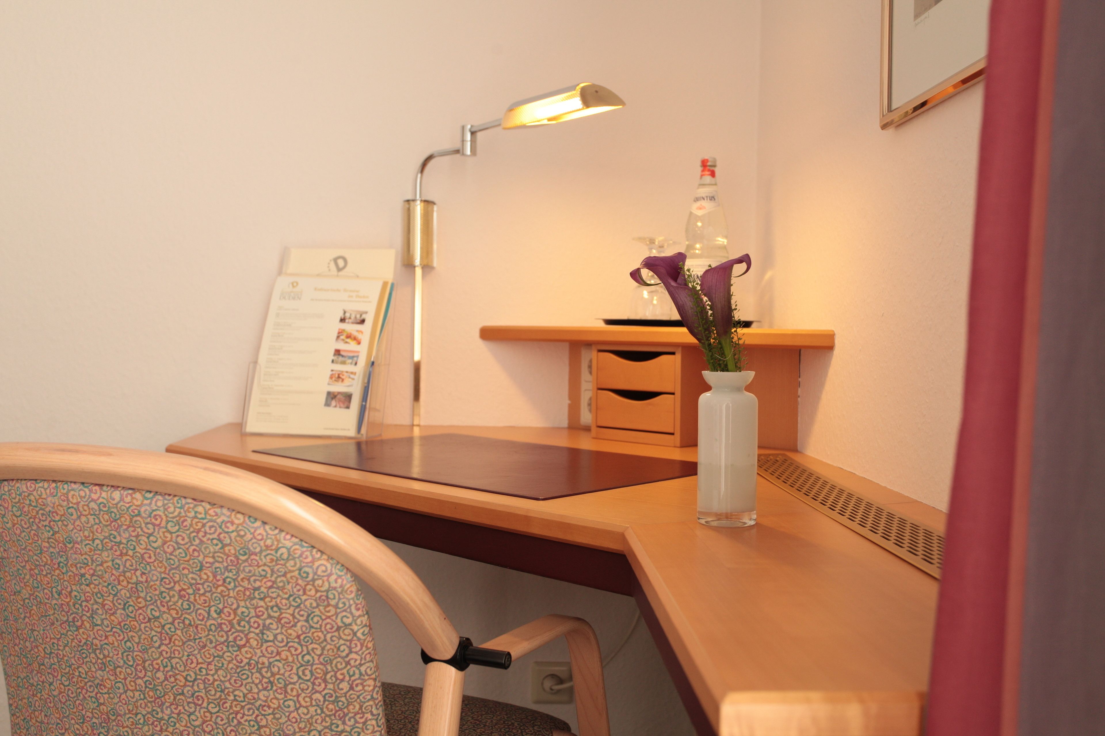 Een hotelkamer met een houten bureau, een lamp, een vaas met bloemen, een fles en een bord met informatie.