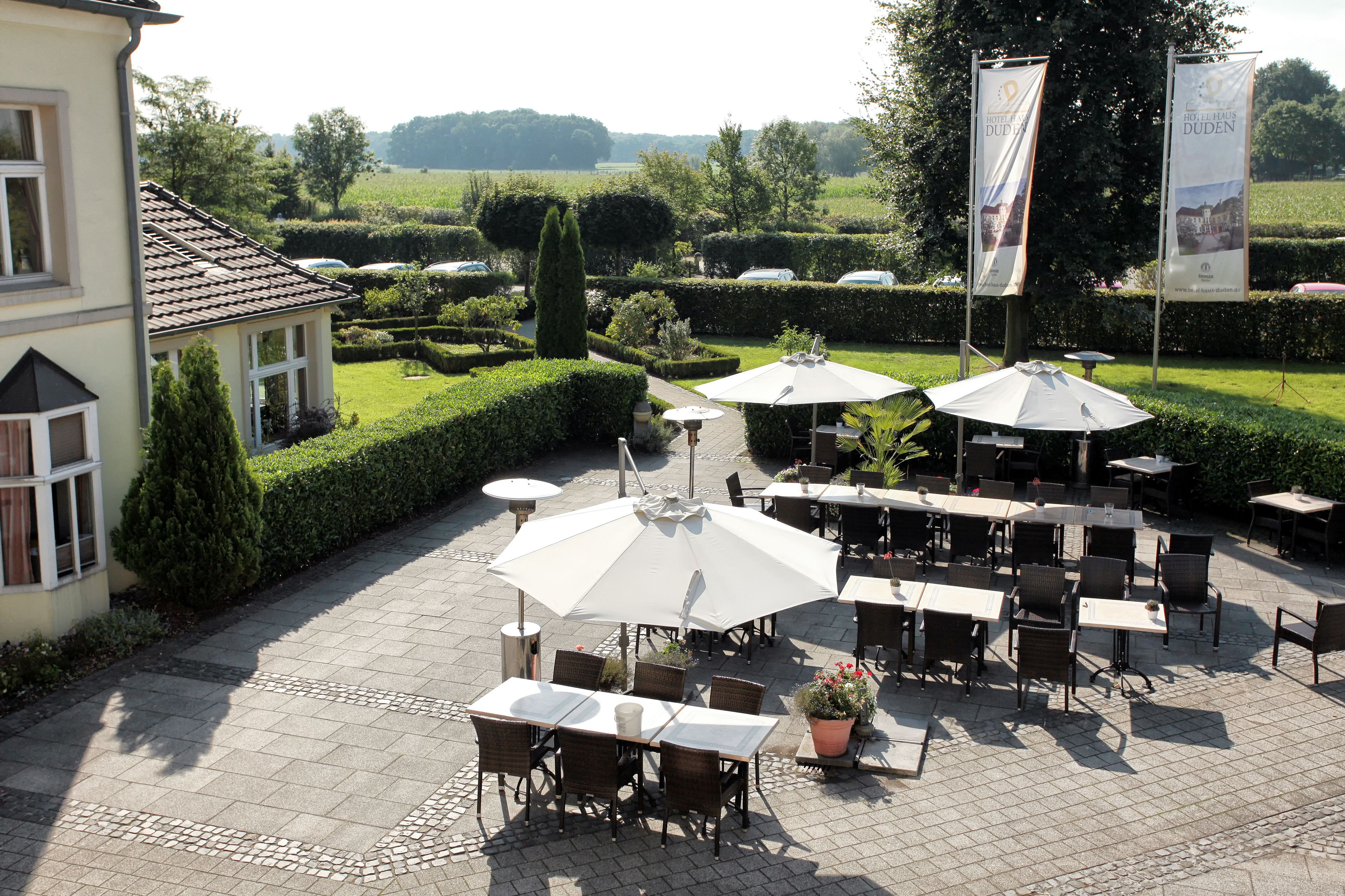 An outdoor patio area with tables and chairs, surrounded by neatly trimmed hedges and a view of a landscaped garden.