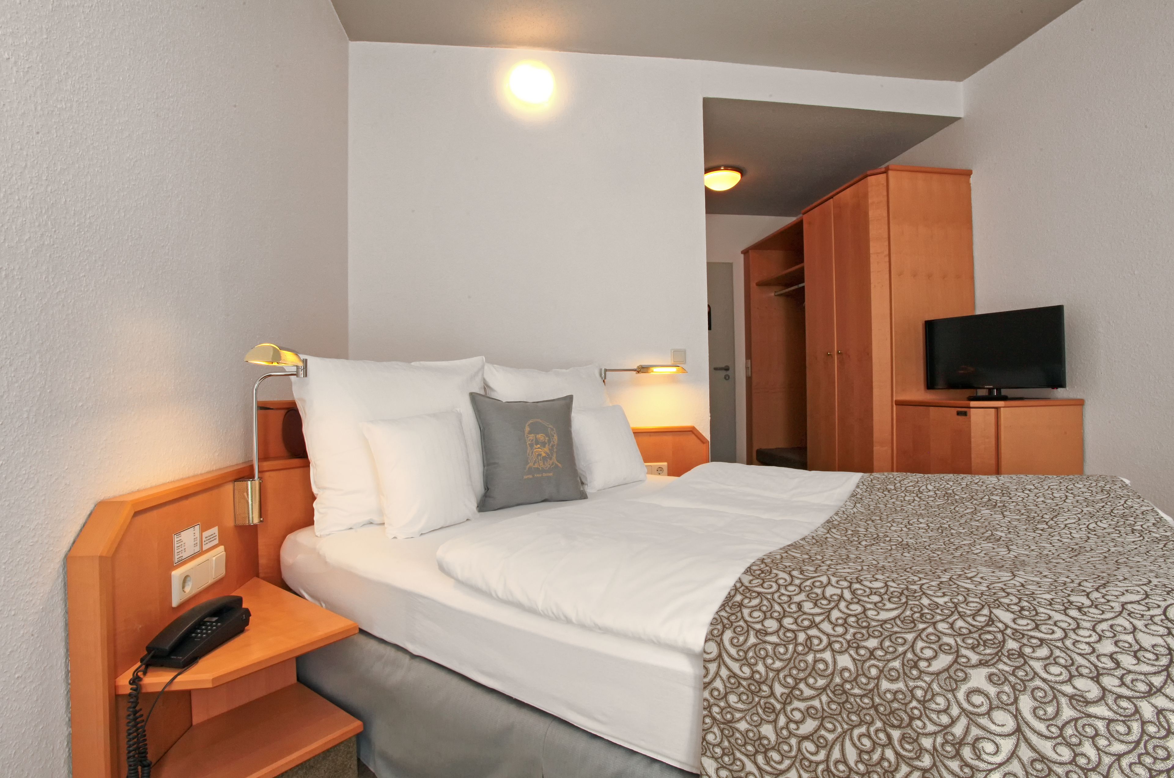 Habitación de hotel con cama, lámparas de noche, televisión, armario de madera y teléfono en la mesita de noche.