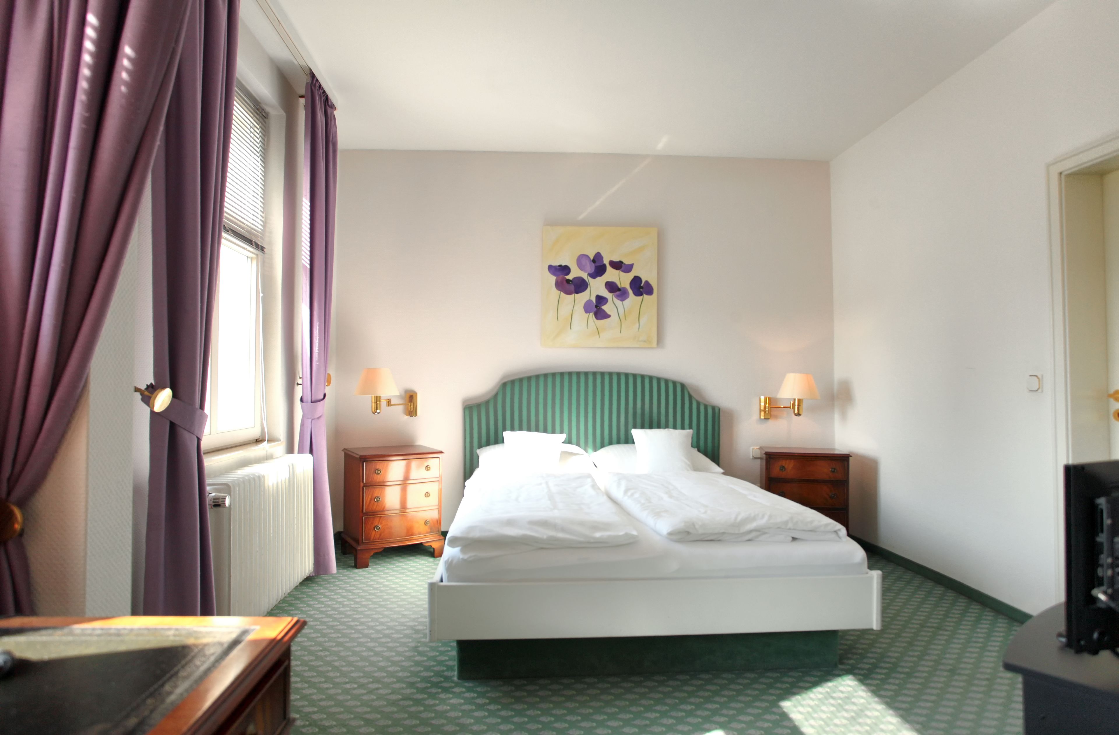 A hotel room with a bed, two bedside tables, a nightstand, a window with purple curtains, a wall-mounted lamp, and a painting.