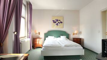 Habitación de hotel con cama, dos mesitas de noche, una lámpara de pared y un cuadro, además de una ventana con cortinas moradas.