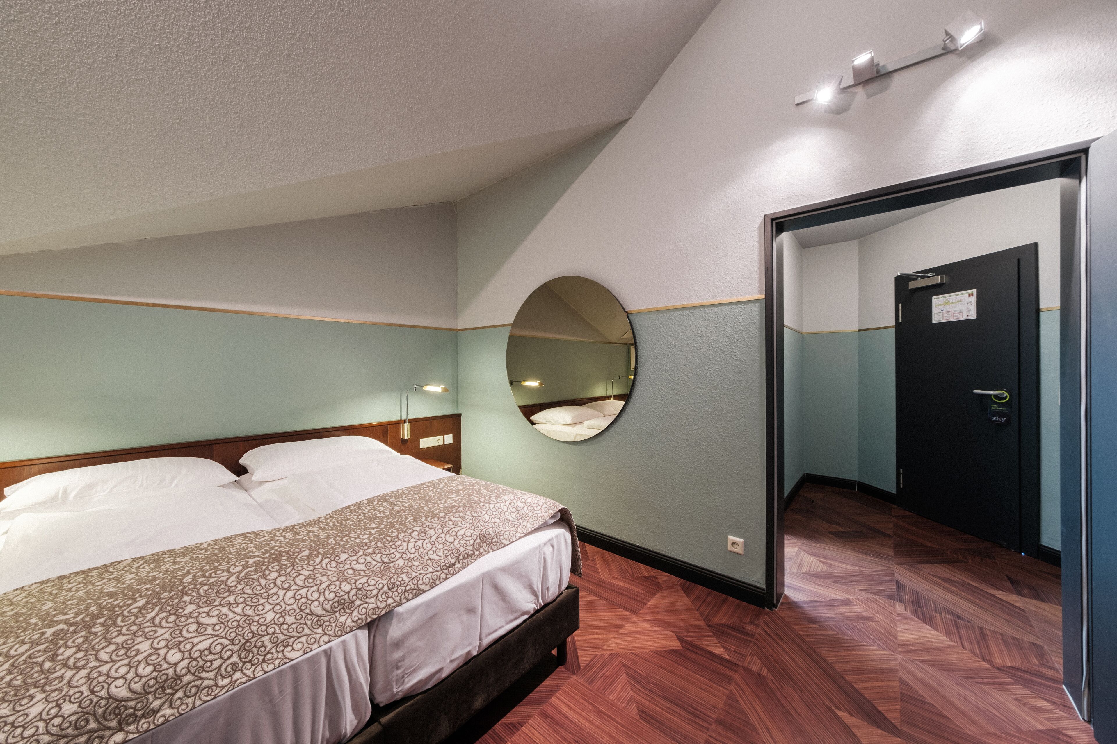 Ein Hotelzimmer mit einem Bett, einem runden Spiegel an der Wand, einer Tür und einem Holzboden.