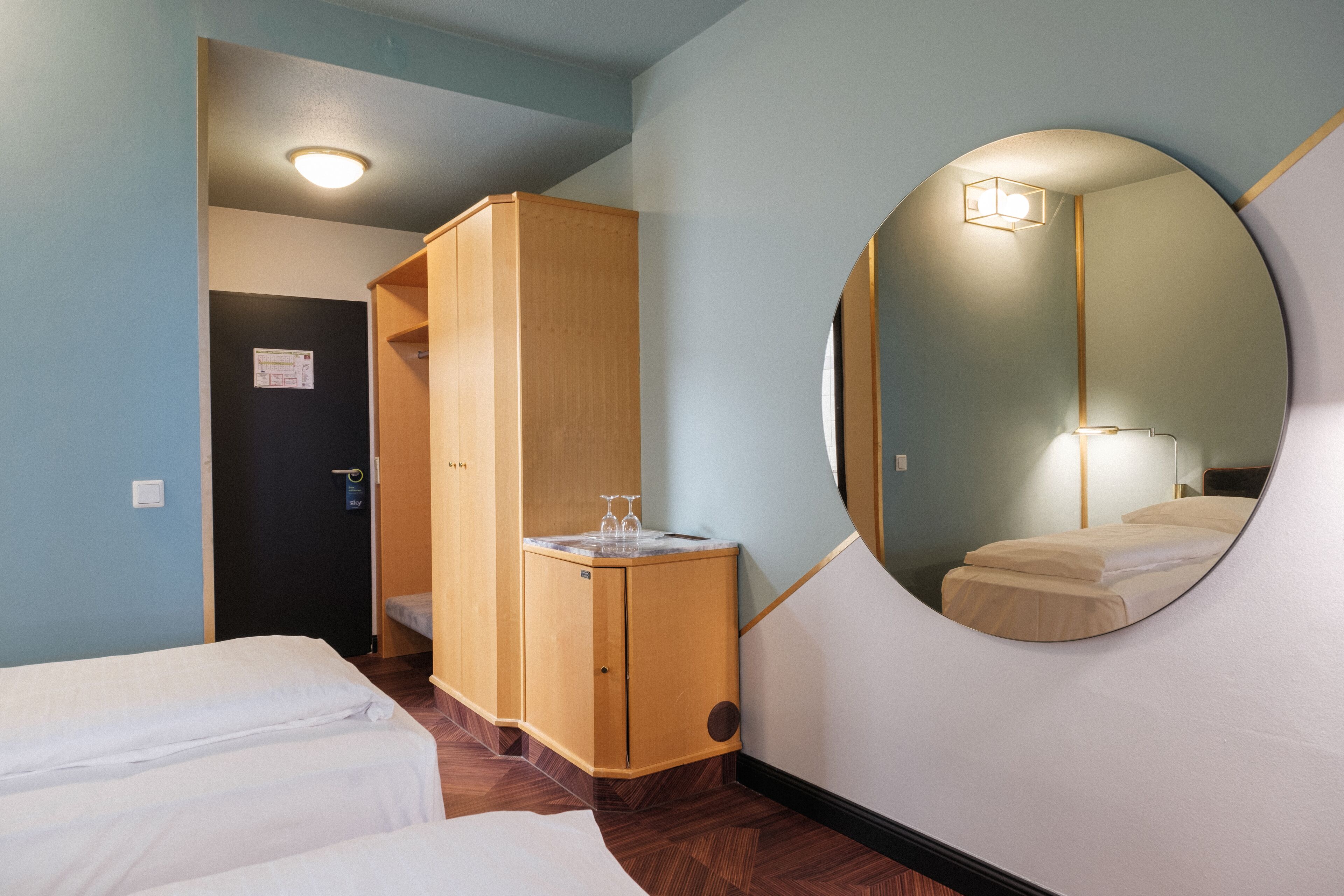 Ein Hotelzimmer mit zwei Einzelbetten, einem Holzschrank, einem runden Spiegel und einem kleinen Tisch mit einem Glas.