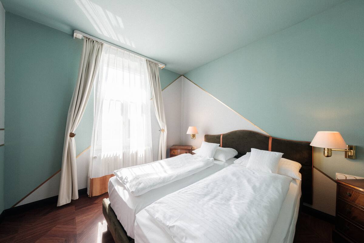 Ein Hotelzimmer mit zwei Betten, einem Nachttisch mit Lampe und einem Fenster mit Vorhängen.