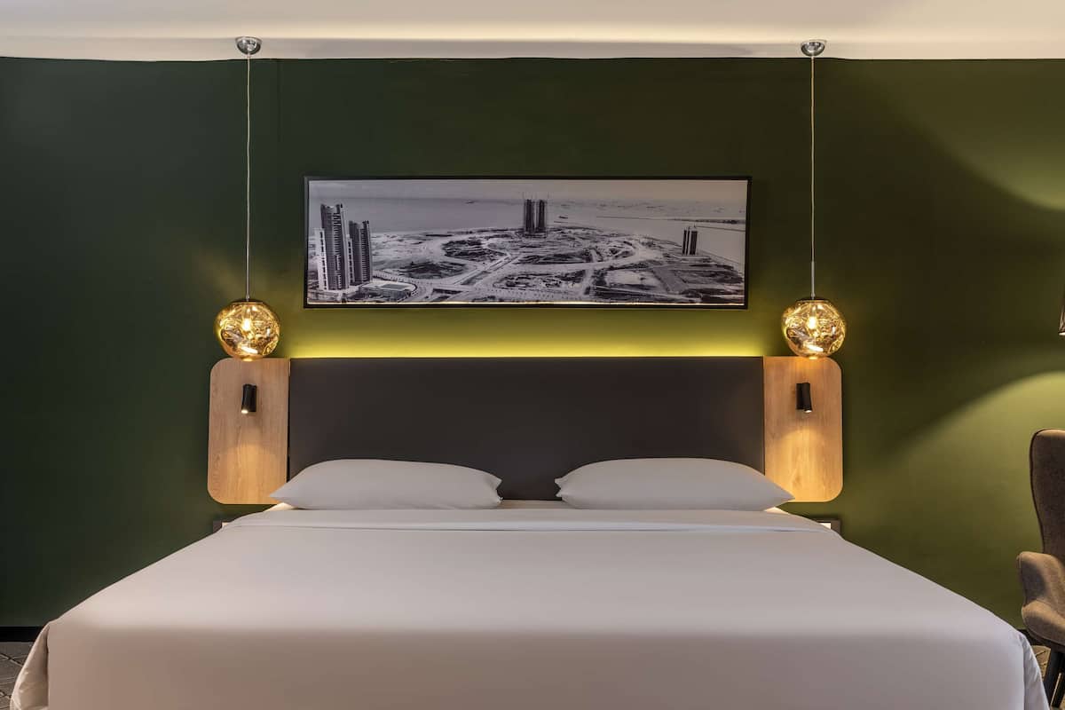 A hotel room with a bed, two wall-mounted lamps, a framed cityscape picture, and a chair.