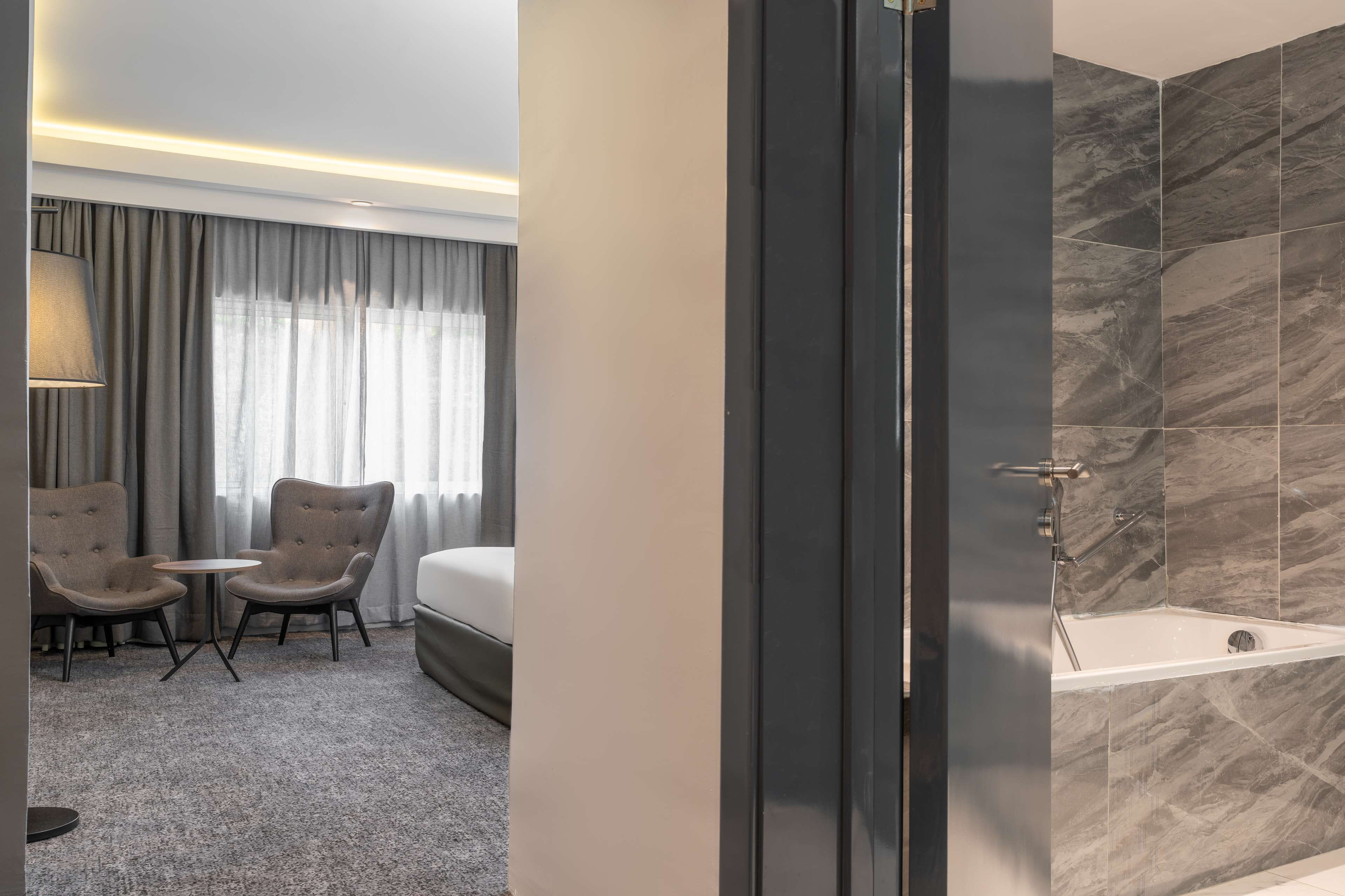 A hotel room with a bed, two armchairs, a small table, a lamp, and a shower area with marble tiles.