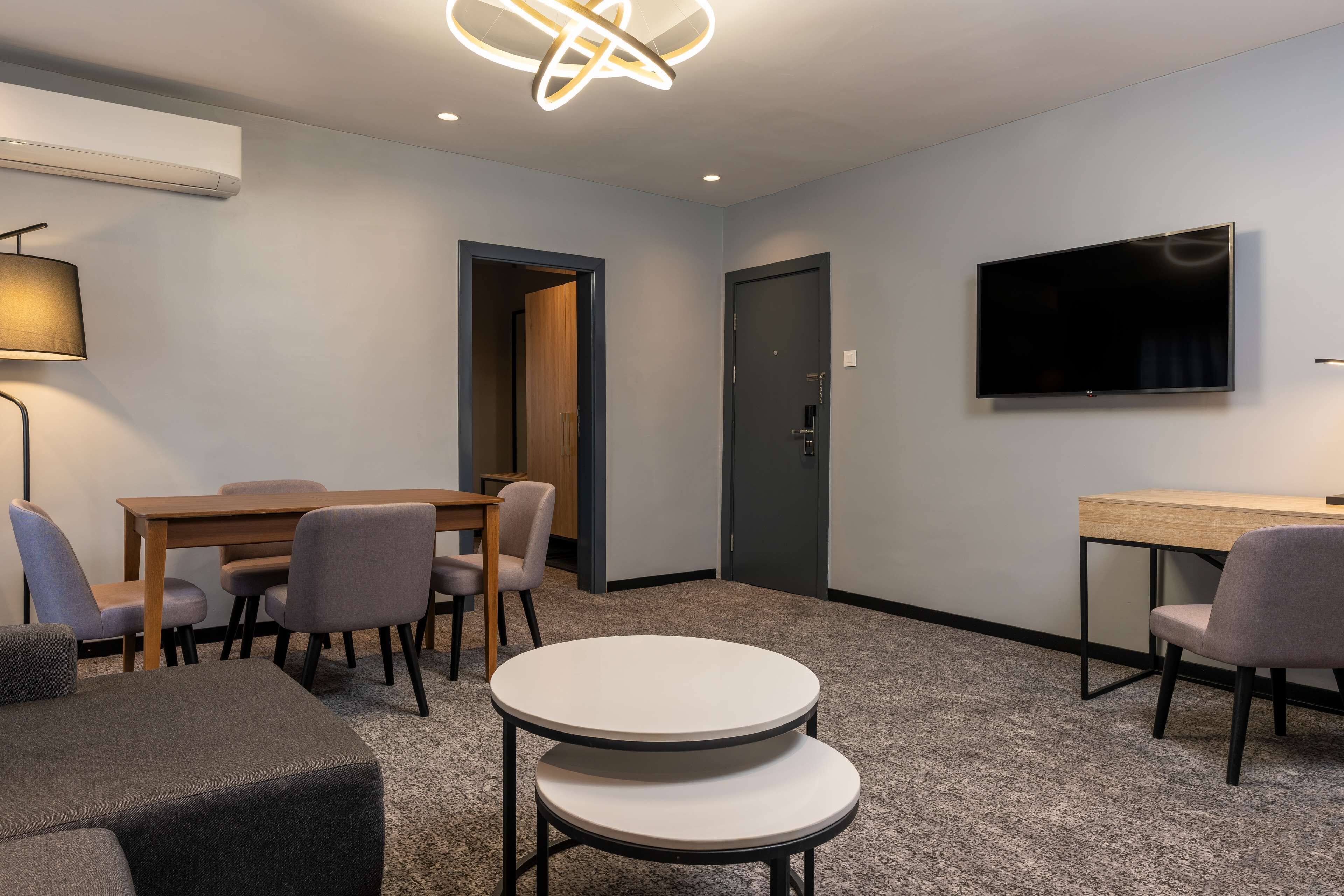 A modern hotel room with a round table, a sofa, a TV mounted on the wall, and a door leading to another room.