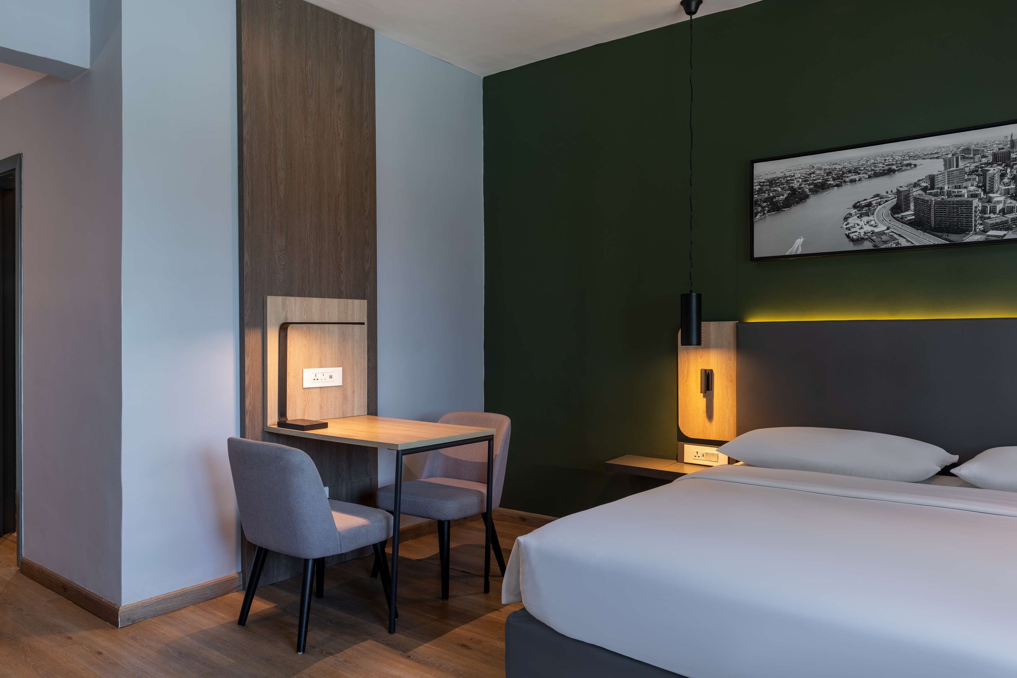 A hotel room with a bed, a desk with two chairs, a nightstand with a lamp, and a cityscape painting on the wall.