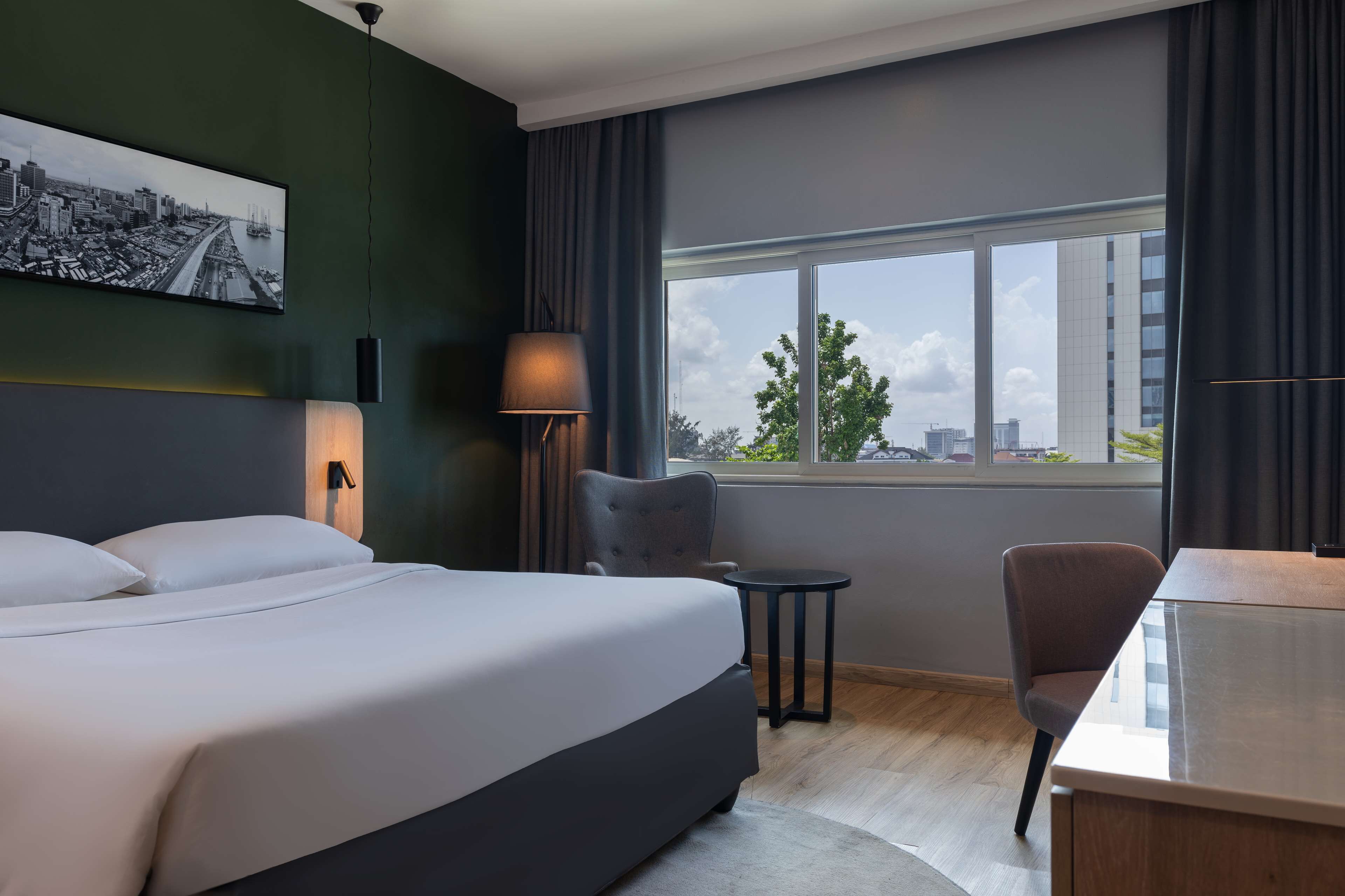 A modern hotel room with a large bed, a desk, a chair, a small table, a lamp, and a cityscape view through the window.