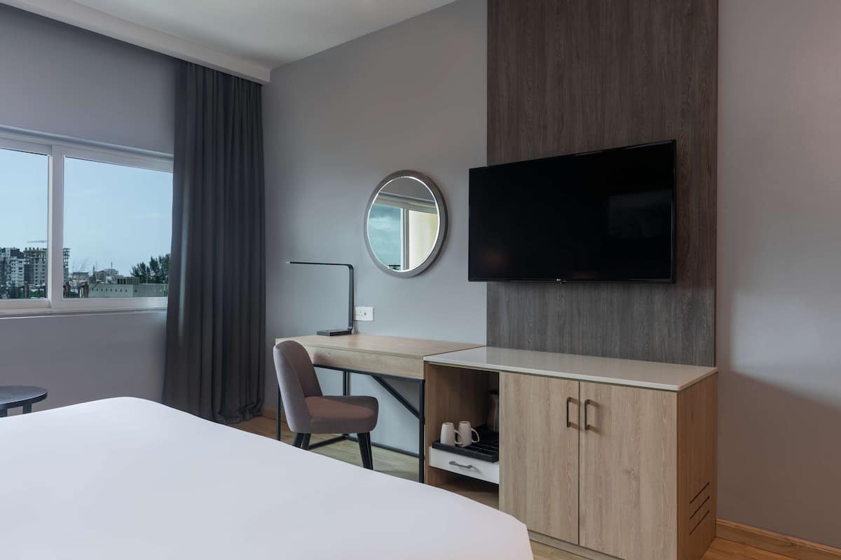 A hotel room with a bed, a desk, a chair, a TV, a mirror, and a view of buildings outside the window.