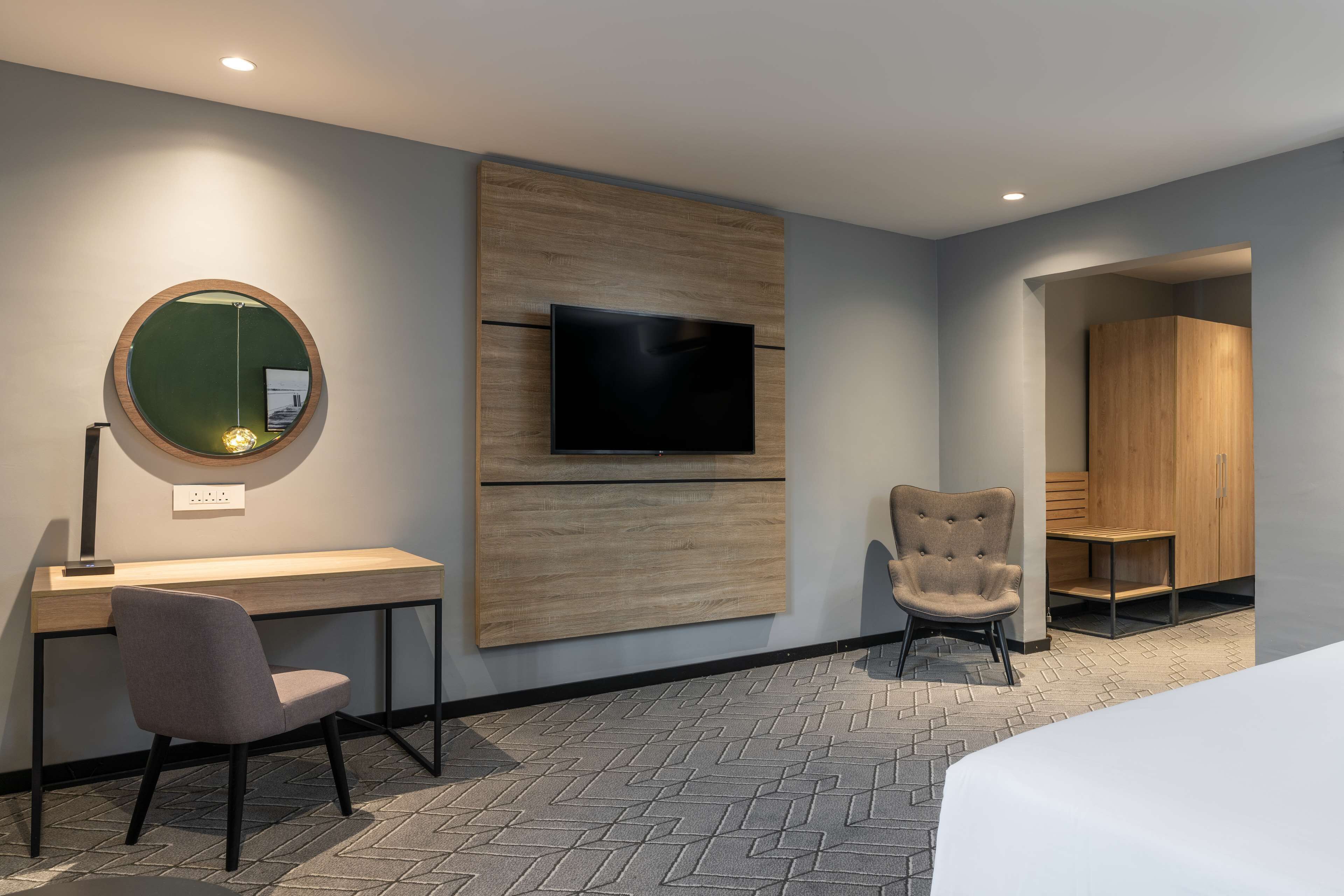 A modern hotel room with a wooden desk, a round mirror, a flat-screen TV, a comfortable armchair, and a wardrobe.