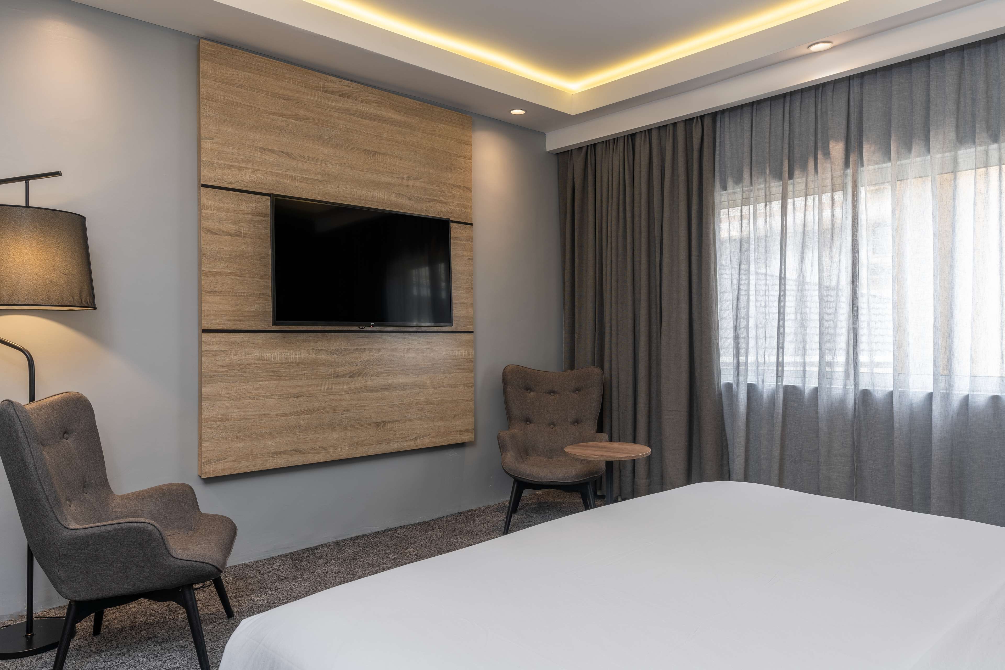 A hotel room with a large bed, a TV mounted on the wall, two armchairs, a small table, and a lamp.