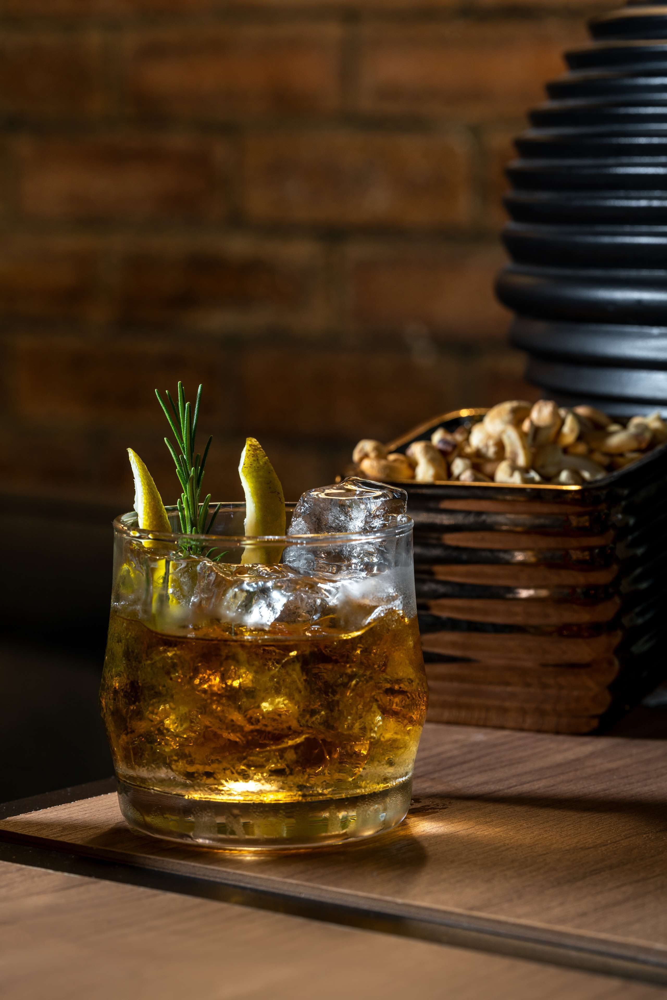 A glass of amber-colored beverage with ice cubes and lemon slices, garnished with rosemary, placed on a wooden surface.