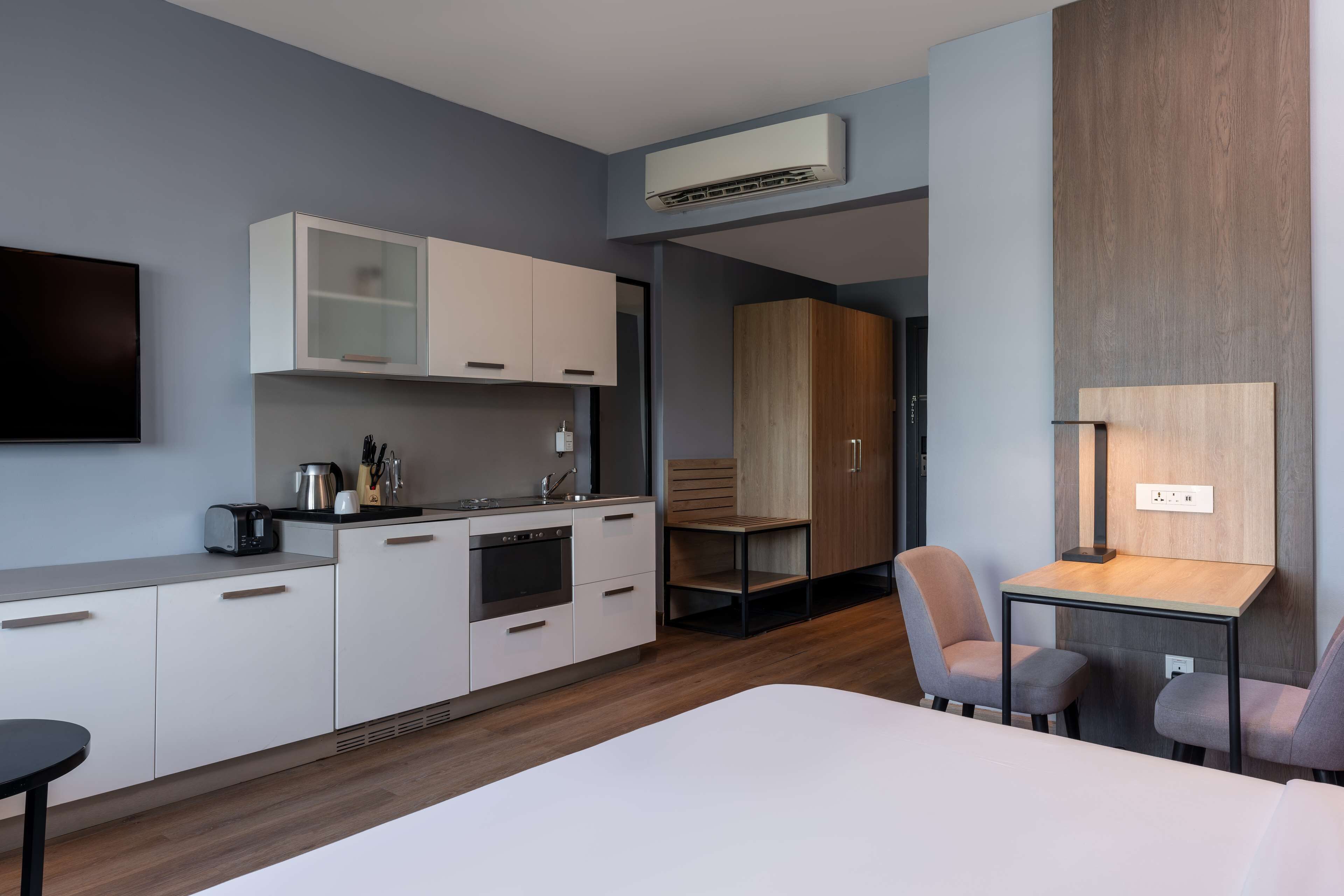 A modern hotel room with a kitchenette, a flat-screen TV, a bed, and a small table with chairs.