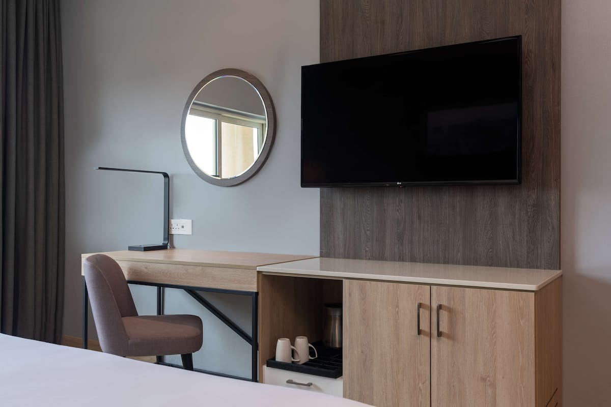 A hotel room with a bed, a desk with a chair, a television mounted on the wall, a round mirror, and a wooden cabinet with a shelf.