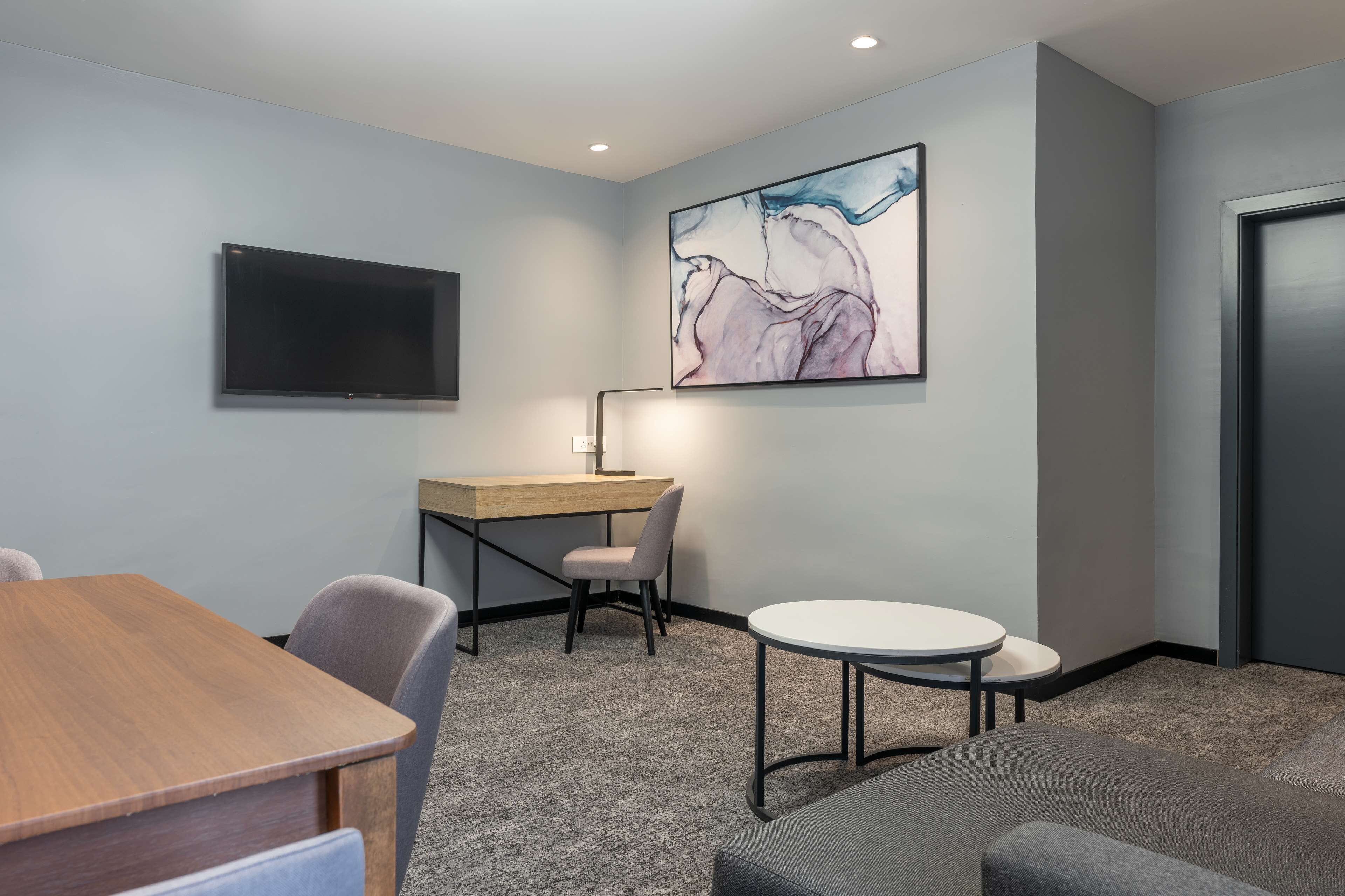 Een moderne vergaderruimte met een flatscreen televisie, een bureau, een stoel, een ronde tafel en een groot abstract schilderij aan de muur.
