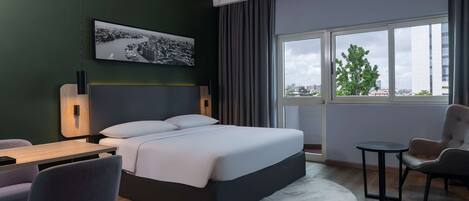 A modern hotel room with a large bed, a desk, a chair, and a view of the city through large windows.