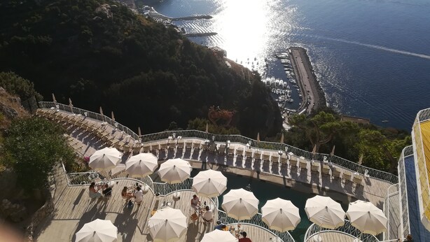 A seaside terrace with white umbrellas and tables, overlooking a coastal area with buildings and boats.