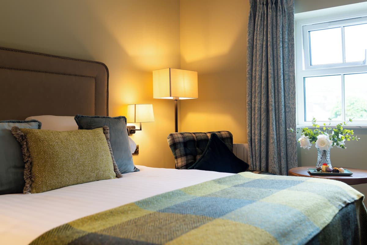 A hotel room with a bed, two bedside lamps, a chair, a small table with a vase of flowers, and a window with curtains.