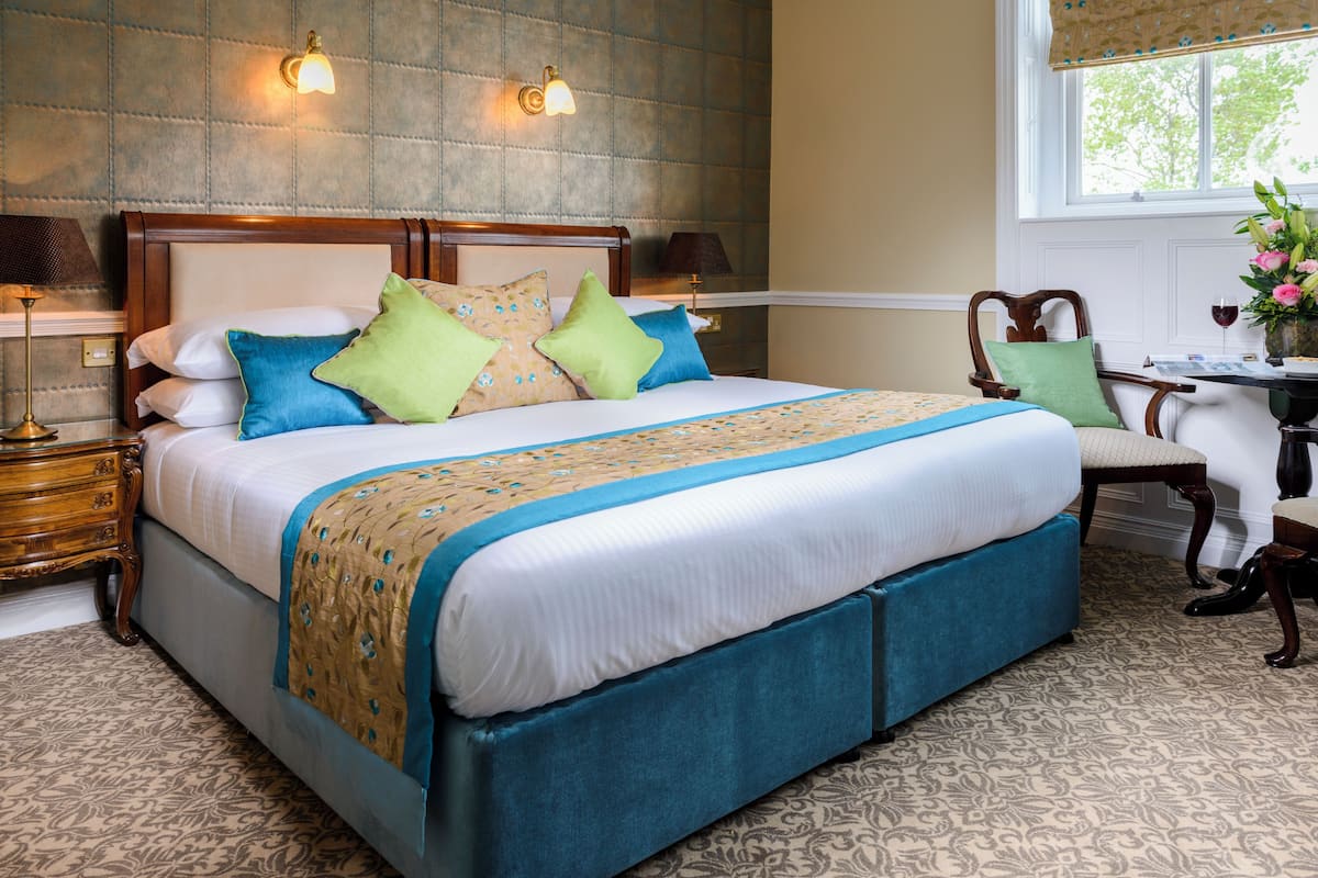 A neatly made bed with a blue and beige bedspread, a wooden headboard, and multiple pillows. There are two bedside lamps and a small table with a vase of flowers.