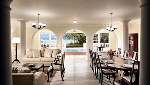 A spacious dining area with a view of a pool and garden, featuring a long table set for dining, comfortable seating, and elegant lighting.