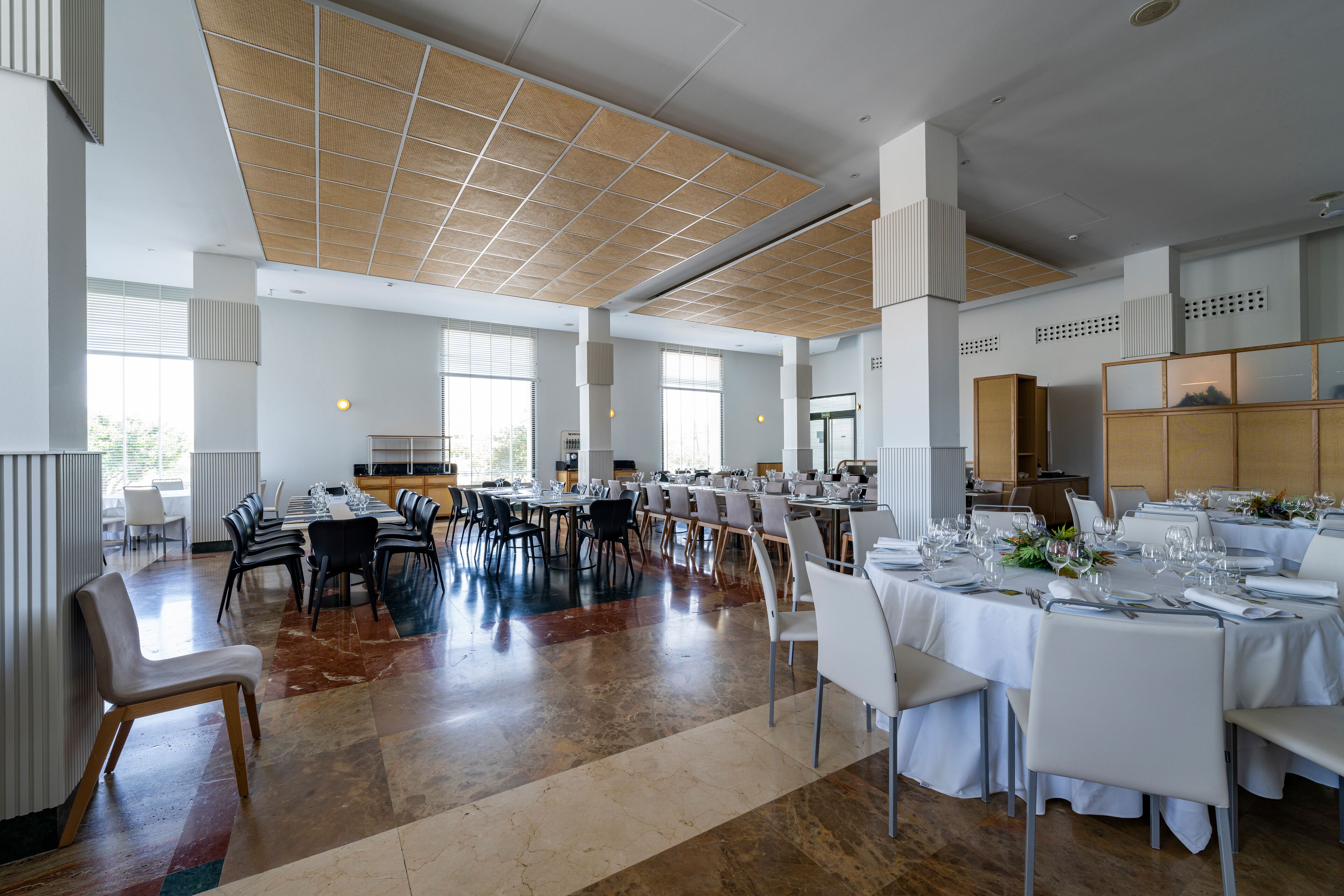A spacious dining area with high ceilings and large windows, featuring multiple tables set for dining.